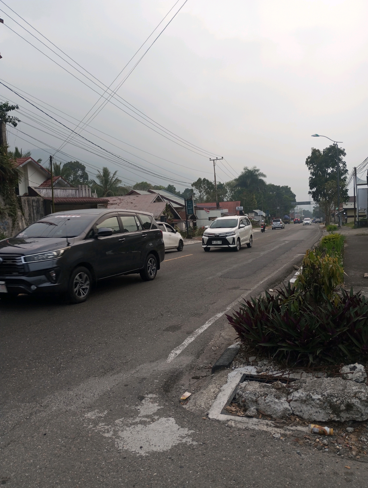The image depicts a street scene in Padang Panjang, Indonesia. The sky is overcast, suggesting a cloudy day or haze. The road is paved, with a white line marking the lane. Several vehicles are visible: a dark-colored SUV in the foreground, a white car in the middle ground, and other cars and a motorcycle further down the road. Buildings, including houses with reddish-brown roofs, are situated along the left side of the street. Trees and utility poles are also present, with numerous overhead wires crisscrossing the sky. The right side of the road features a sidewalk with decorative plants and some damaged concrete infrastructure. Litter, including a plastic bottle and a crushed can, is scattered on the roadside. The overall impression is of a typical urban or semi-urban environment on a somewhat hazy day.