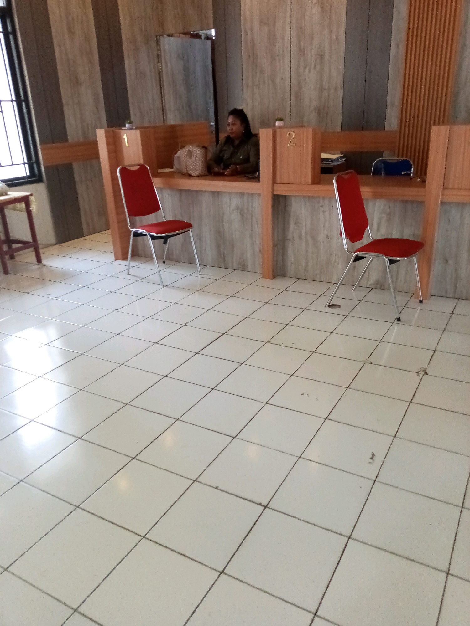 The image shows an interior scene, likely a bank or government office, with a tiled floor and wood-paneled walls. In the background, a woman is seated behind a counter, appearing to be an employee. Two red chairs with silver frames are positioned in front of the counter, suggesting a waiting area. Numbers "1" and "2" are visible on wooden dividers at the counter, indicating service points. The overall impression is that of an organized and functional service space. This scene is located in Bitung, Indonesia.