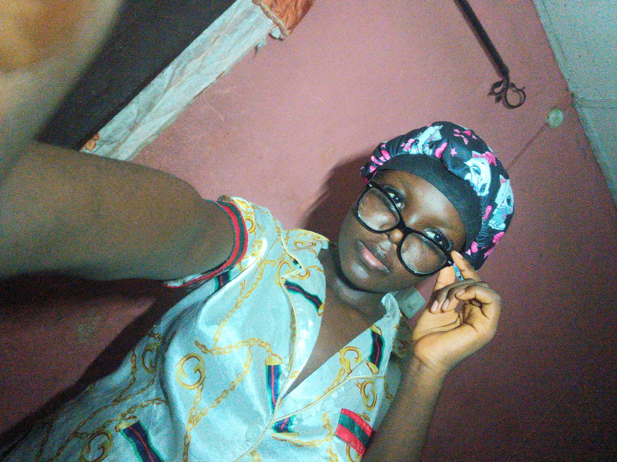 An indoor, self-portrait-style photograph features a young person, likely female, with dark skin, looking directly at the camera. The subject is wearing large, round, black-rimmed eyeglasses, which they are casually adjusting with their left hand. A black sleep bonnet, adorned with a repeating pattern of white skulls and pink bows, covers their hair. They are dressed in a light-colored, possibly satin, pajama top featuring an intricate design of gold chains interwoven with red, green, and dark striped bands. The right arm of the subject is partially visible on the left side of the frame, indicating they are holding the device used to capture the image.
The background consists of a plain, warm-toned wall, appearing in a shade of pinkish-brown or terracotta. In the upper right corner, a dark, thin, curved object, possibly a hook or clothes hanger, is visible. The upper left part of the frame shows an indistinct, darker area, possibly a shadow or part of another object, next to a lighter, fabric-like surface.
The lighting in the image is somewhat dim, resulting in a noticeable grainy texture across the photograph. The subject maintains a neutral to slightly pleasant expression, with a subtle hint of a smile. The scene conveys a casual, personal moment within what appears to be a private room or home in Onitsha, Nigeria. There is no visible text in the image.