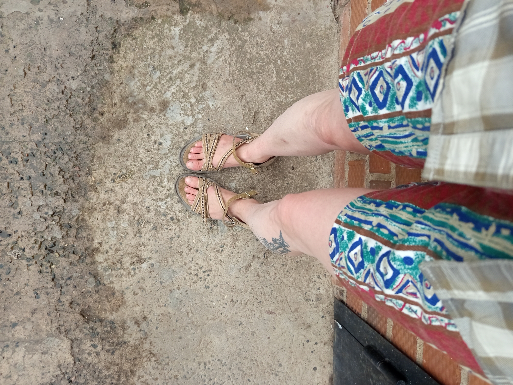 This image shows a close-up, high-angle view of a person's legs and feet, standing on a concrete surface. The person is wearing tan-colored sandals with fabric straps. They are also wearing shorts with a patterned fabric that includes geometric designs in blue, white, and green, bordered by red and brown. The background is a rough, textured concrete surface, possibly an outdoor patio or walkway, with some small stones and darker patches suggesting moisture or dirt. The lighting appears to be natural, indicating daytime. A faint tattoo is visible on the person's left shin. The overall impression is casual, as if someone is posing for a photograph outdoors.