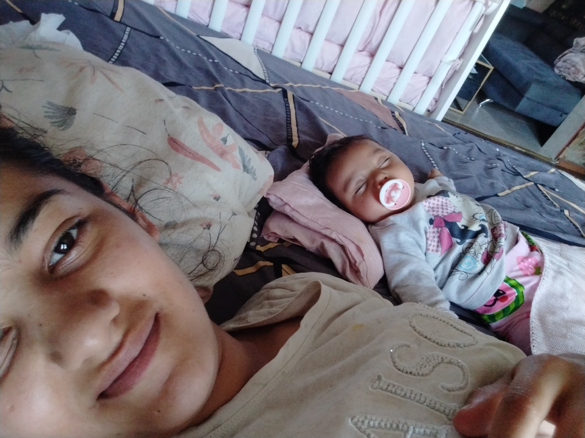 A selfie of a woman with a baby, likely in a domestic setting. The woman, located in the lower left foreground, is partially visible with a focus on her face; she is smiling. Next to her lies an infant with a pacifier in its mouth, asleep on a pillow. They are both lying on a bed with patterned bedding. Adjacent to the bed is a baby crib with pink cushioned sides. The background includes a glimpse into another room, revealing a sofa and other furnishings. The exact location is not discernible from the image.