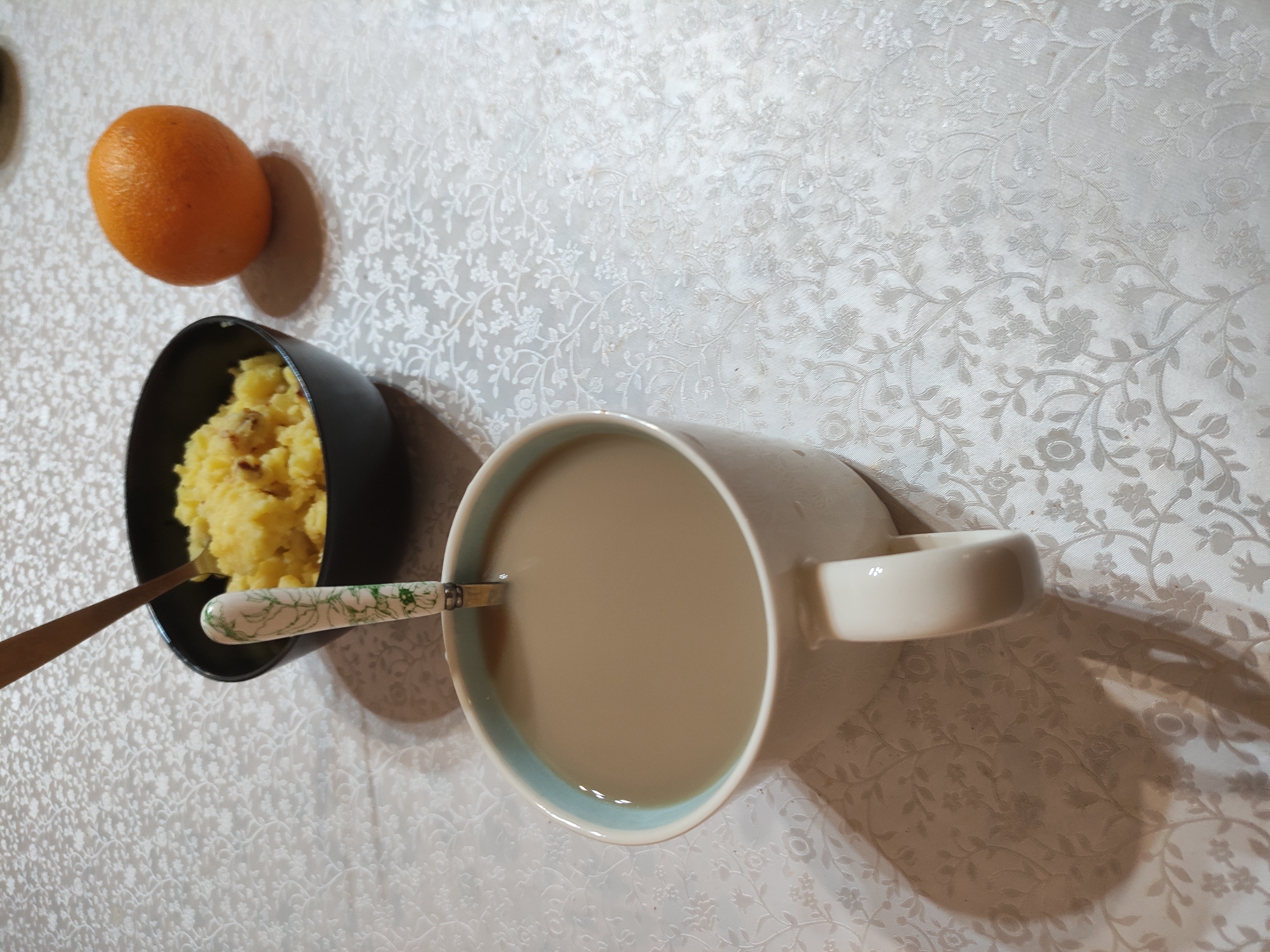 On a white tablecloth with a floral pattern, a meal is arranged. There is an orange, a small black bowl of mashed potatoes with a golden spoon protruding from it, and a large mug filled with a tan beverage, a spoon with a green floral pattern rests inside. The shadows suggest a light source from above. The location is Unknown, Unknown.