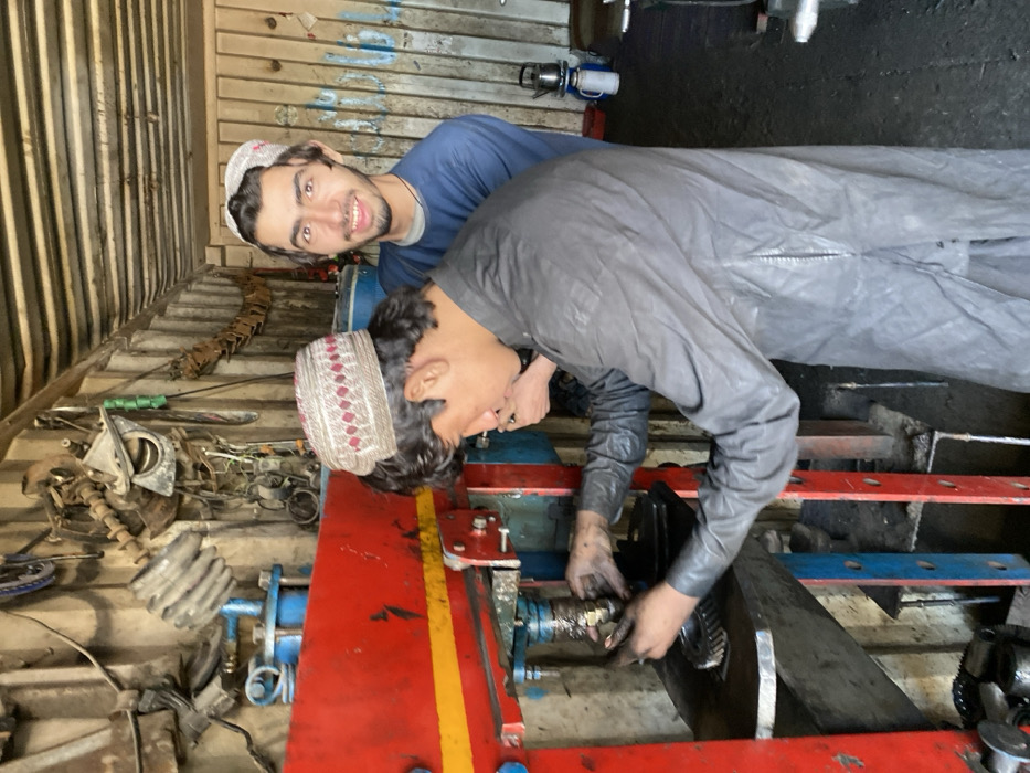 Two young males are depicted within what appears to be a metalworking or mechanic's workshop, likely located in an industrial or commercial area of Kandahar, Afghanistan.
The primary subject, positioned in the foreground and facing left, is a male with dark hair wearing a light-colored, intricately patterned skullcap and a dark-grey, long-sleeved tunic over trousers. His hands, soiled with grease, are actively engaged in manipulating metal components within a large, red hydraulic press. A metal gear is visible on the press's platform below his hands.
Behind him, partially obscured, stands a second male, also wearing a patterned skullcap and a blue long-sleeved shirt. He is looking directly at the camera with a slight smile. His right hand is visible, positioned near the first male's back, but his specific action or interaction is unclear due to the partial view.
The workshop environment features corrugated metal walls, indicative of a repurposed container or simple construction. The walls are cluttered with various metal parts, tools, and machinery components hanging or stored haphazardly. A blue barrel or container is visible in the background, along with a kettle and cups on a surface, suggesting a break area. The floor appears dark and potentially unpaved or heavily soiled. Lighting is bright, suggesting natural light from an unseen opening.