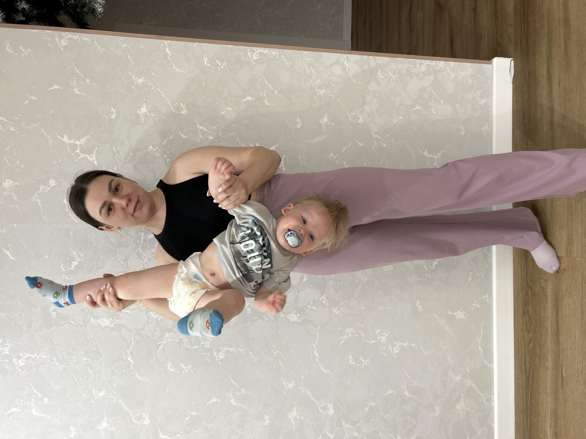 The image depicts a woman lying on a light-colored, possibly marble-patterned surface with wood flooring visible on the right side of the frame. The woman is wearing a black sleeveless top and lilac-colored pants. She is holding a baby with blond hair and a pacifier in his mouth. The baby is wearing a gray shirt and a diaper, and socks. The location is Chaikovsky, Russia. A Christmas tree is partially visible in the upper left corner.