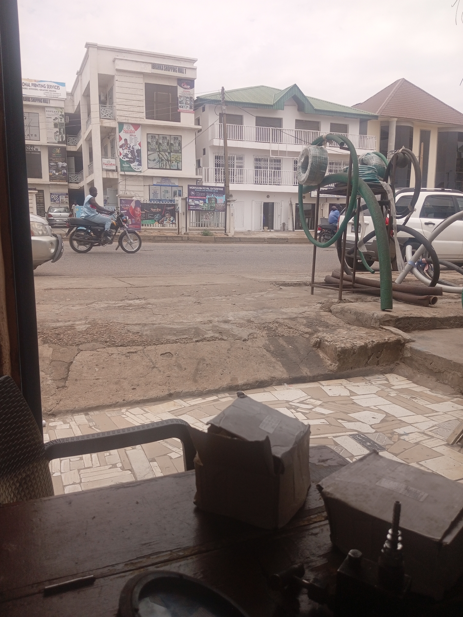 The image depicts a street scene in Sardauna, Nigeria, with a focus on a multi-story commercial building identified as "IBRANKKA SHOPPING MALL 1." In the foreground, a dark wooden table is visible with two cardboard boxes and some tools. The middle ground shows a street with a man riding a motorcycle. To the right, there is a white SUV with various hoses attached to what appears to be equipment. The background features the shopping mall and other buildings, suggesting a commercial area within the city. The sky is overcast.