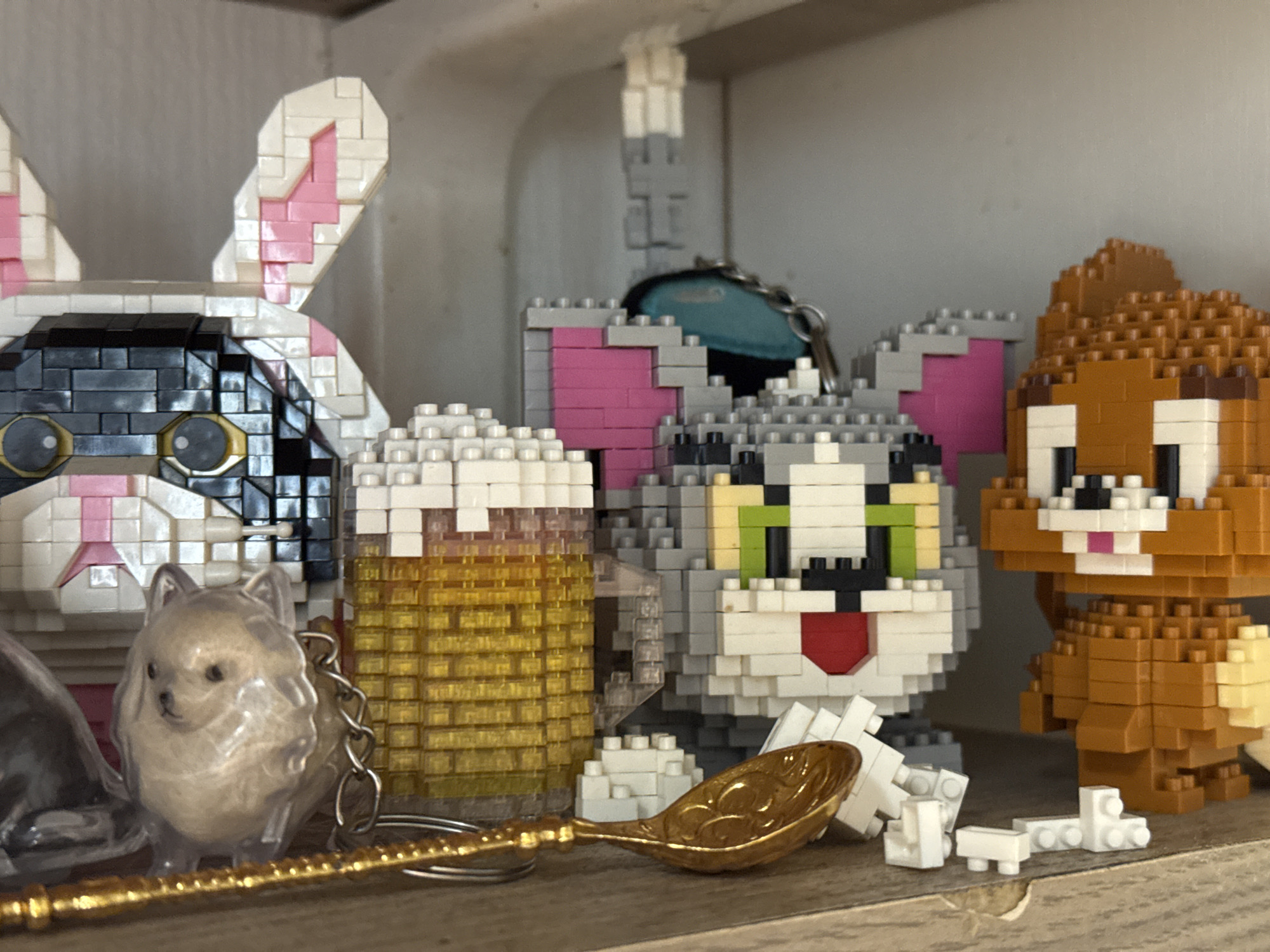 The image shows a collection of toy figures arranged on a shelf, likely indoors, in an unspecified location. From left to right, there is a rabbit made of white, black, and pink blocks. To its right is a transparent dog keychain. Next to that is a golden spoon laying next to a cup made of yellow blocks. Then, a Tom figure from Tom & Jerry made of gray, white, black, pink, green, and red blocks. Lastly, a Jerry figure from Tom & Jerry made of brown, white, and black blocks. The background is out of focus but suggests a neutral-colored wall and shelf environment. There is no evidence to indicate this image originated from Saint Petersburg, Russia.