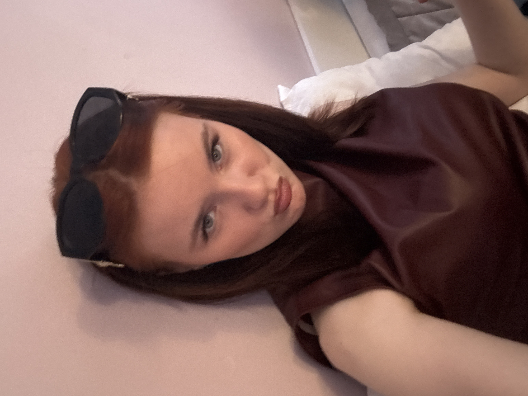 A woman lies on her back on a light-colored surface, presumably a bed, with white pillows visible in the upper right corner. She has auburn hair and is wearing sunglasses on her head. She is also wearing a burgundy shirt. Her face is slightly angled towards the camera, and she has a pursed-lip expression. The background behind her appears to be a light-pink wall. There is no indication of this being Saint Petersburg, Russia.