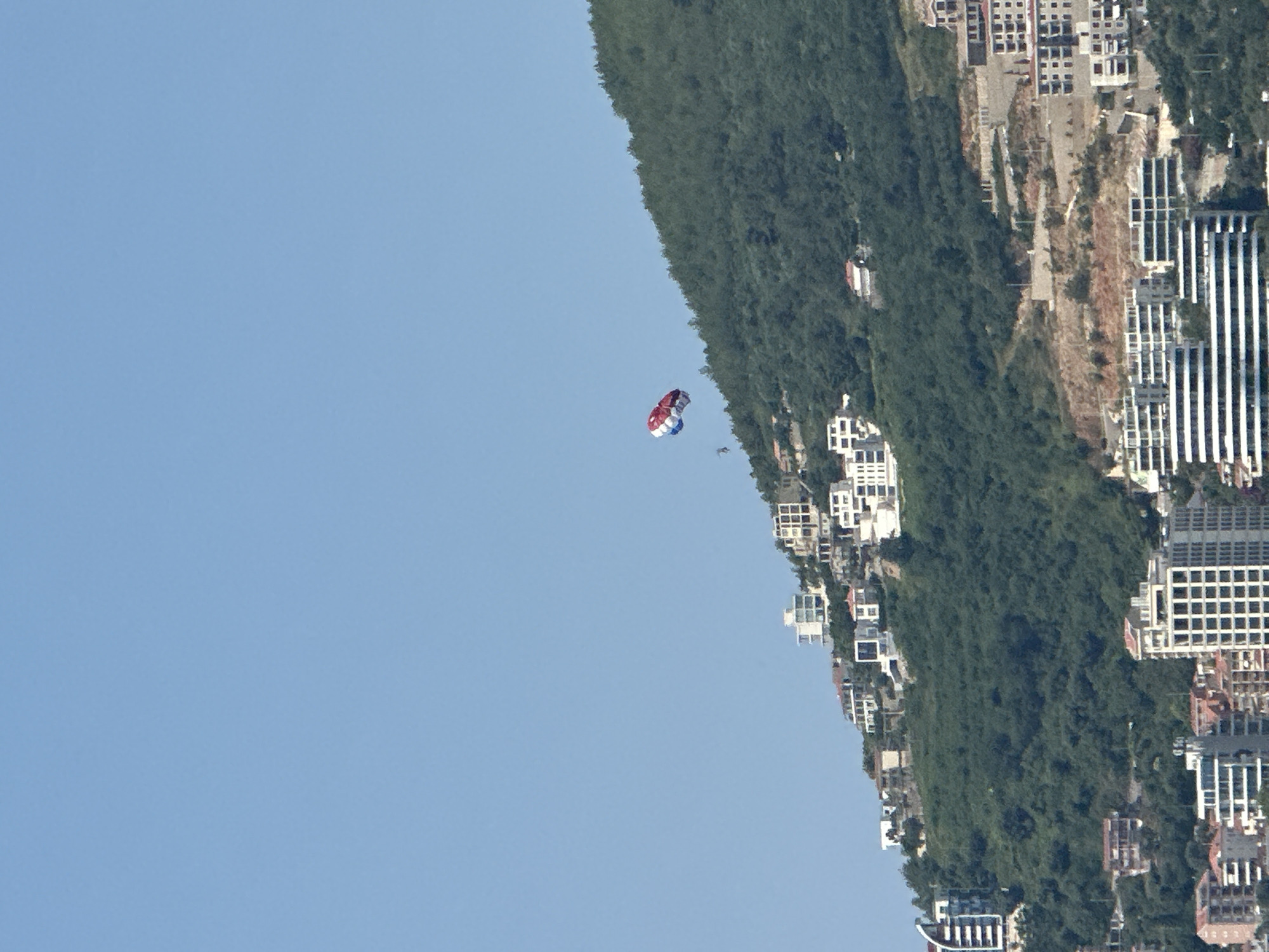 The image shows a parasailer in mid-air against a clear blue sky. The parasail is red and white with some blue accents. Below the parasailer, a green, densely forested hillside slopes downwards, dotted with buildings. These buildings appear to be modern apartments or hotels, clustered together on the steep terrain. Some of the structures have white facades and balconies, while others are taller with grid-like window patterns. The scene is shot from a high angle, looking down onto the hillside and up at the sky. The image is oriented vertically, with the sky on the left and the hillside and buildings on the right. The lighting suggests it is daytime, likely late morning or early afternoon, with the sun casting shadows on the buildings and highlighting the foliage. The overall impression is one of a coastal resort town, possibly Budva, Montenegro, given the context, with recreational activities taking place against a scenic backdrop. There is no visible text in the image.