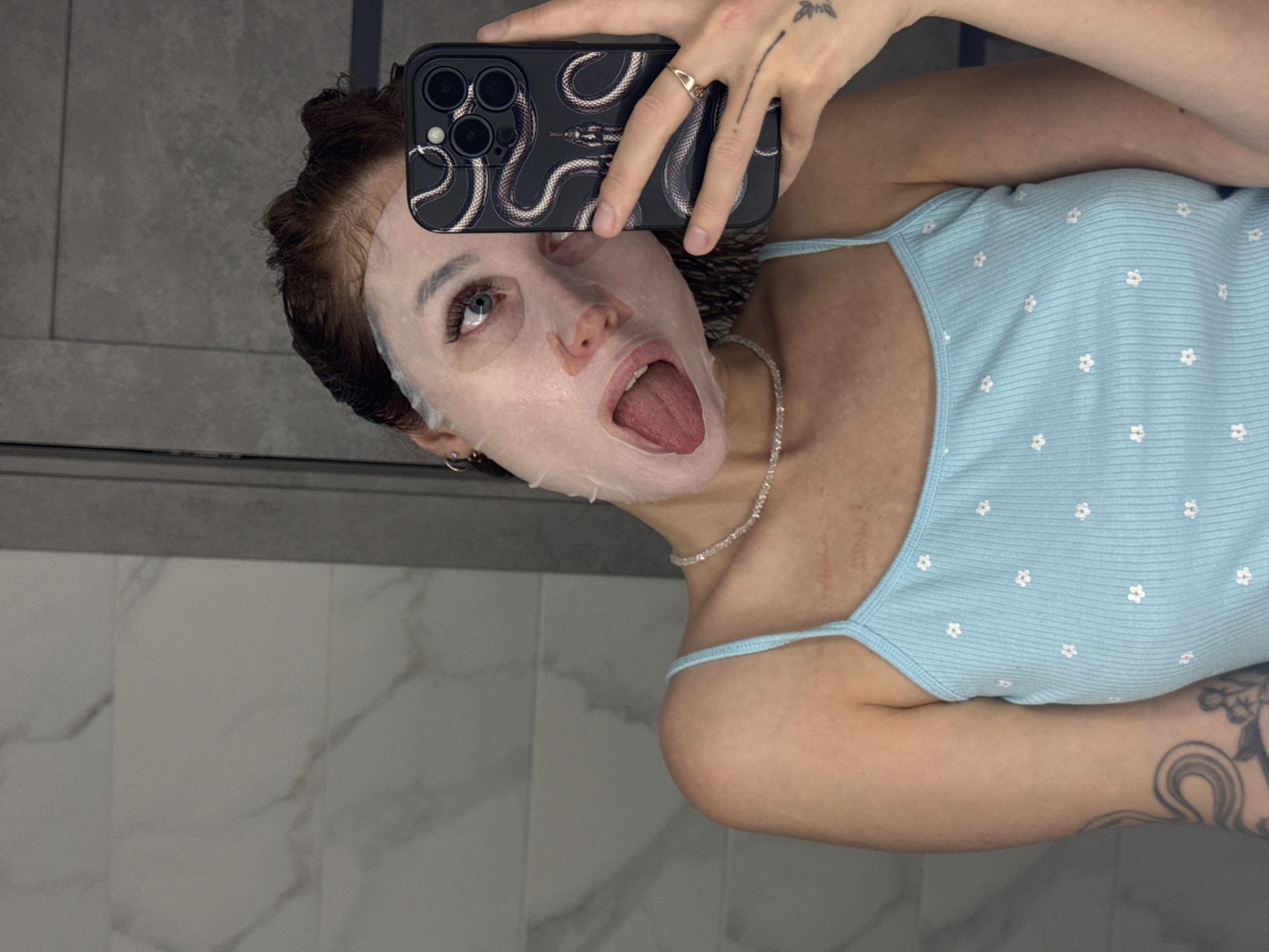 A young woman with her mouth open and tongue out, wearing a white sheet face mask, is taking a selfie in a bathroom. She is wearing a light blue ribbed tank top with a floral pattern and a thin, sparkling necklace. Her hair is dark and pulled back. The background consists of a tiled wall and a gray door. The woman is holding a black smartphone with a snake design in her right hand. She has a ring on her right ring finger and a tattoo on her left arm. The image appears to have been taken in the city of Chaikovsky, Russia, given the context of the prompt.