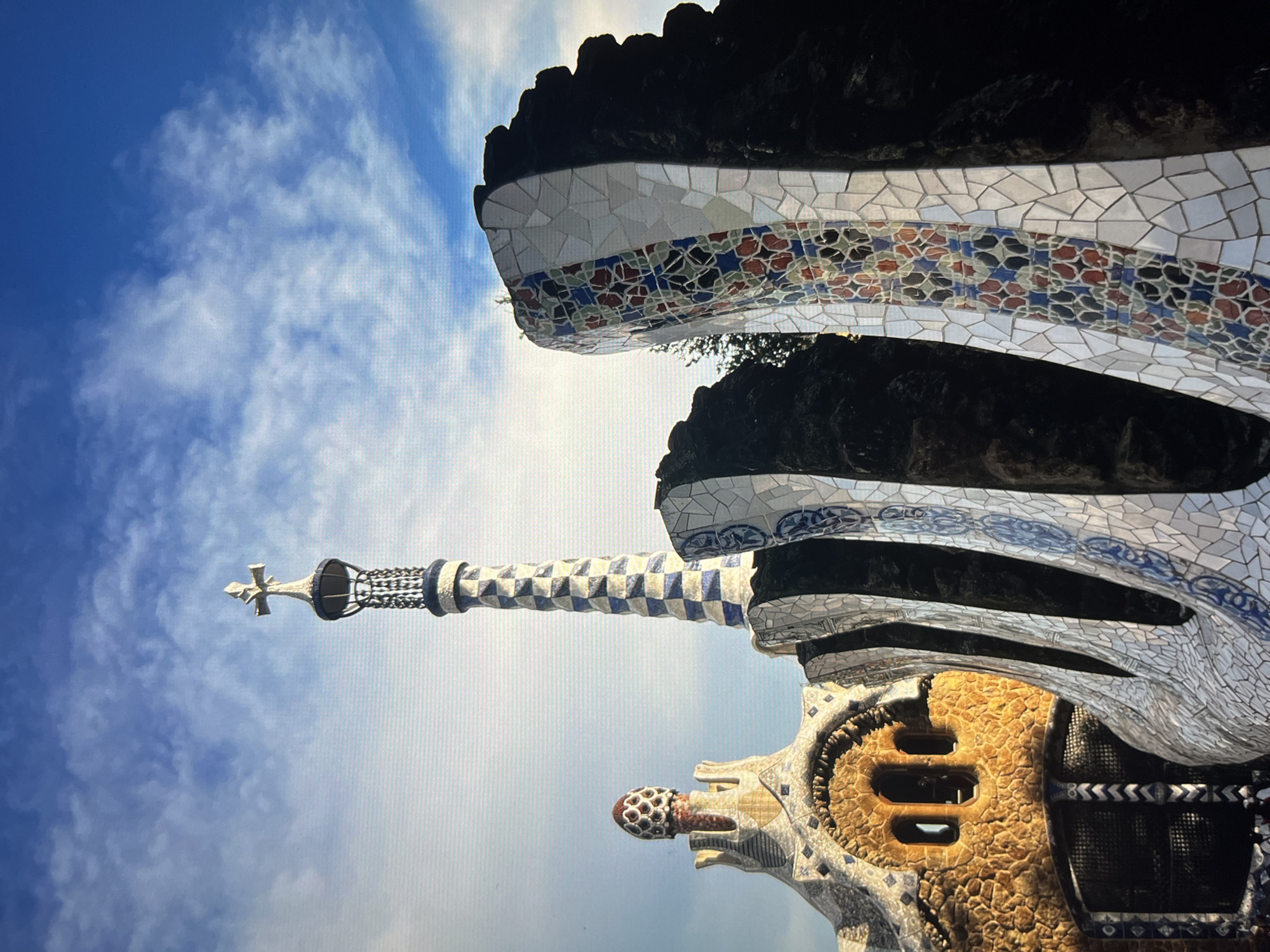 This image depicts a building in Park Güell, Barcelona, Spain, shot from a low angle looking up at the sky. The architectural style is attributed to Antoni Gaudí, known for his distinctive mosaics and organic forms. The building features a tower with a cross at the top, mosaic tiling in various patterns and colors (white, blue, red, orange, black), and structures resembling natural elements. The sky is blue with scattered white clouds, providing a backdrop to the colorful architecture. The overall composition suggests a sunny day.