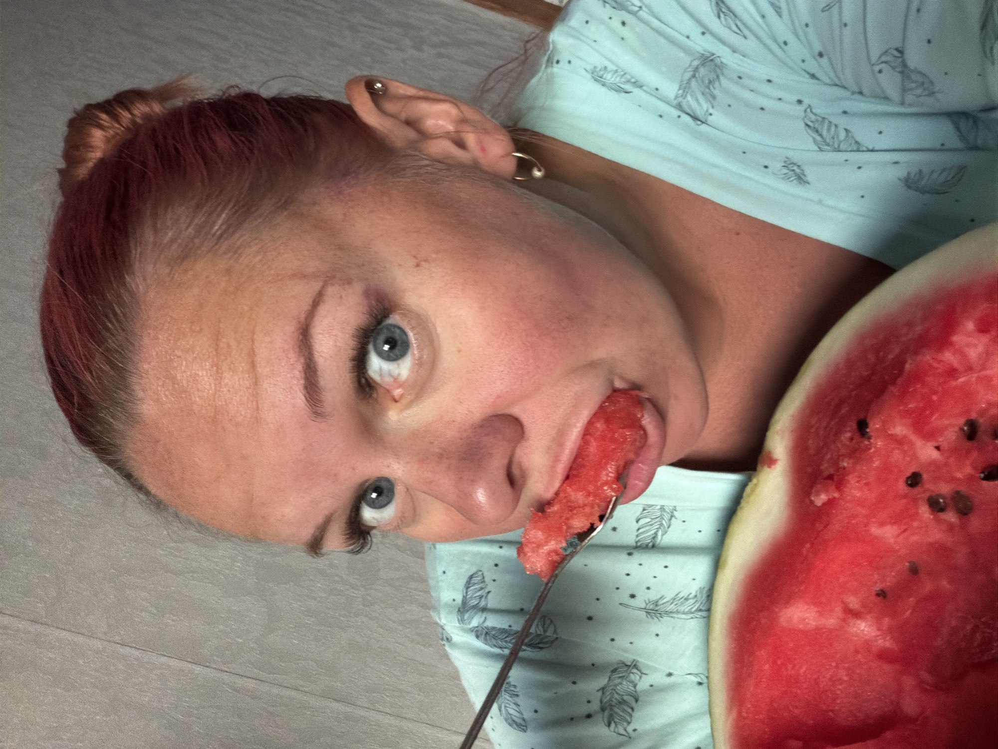 The image shows a close-up of a woman with blue eyes and pinkish-red hair styled in a bun, eating watermelon with a spoon. She is wearing a light blue pajama top with a feather print. The background appears to be a textured gray wall, possibly in a home. There is no indication of the city of Chaikovsky, Russia, in the image.