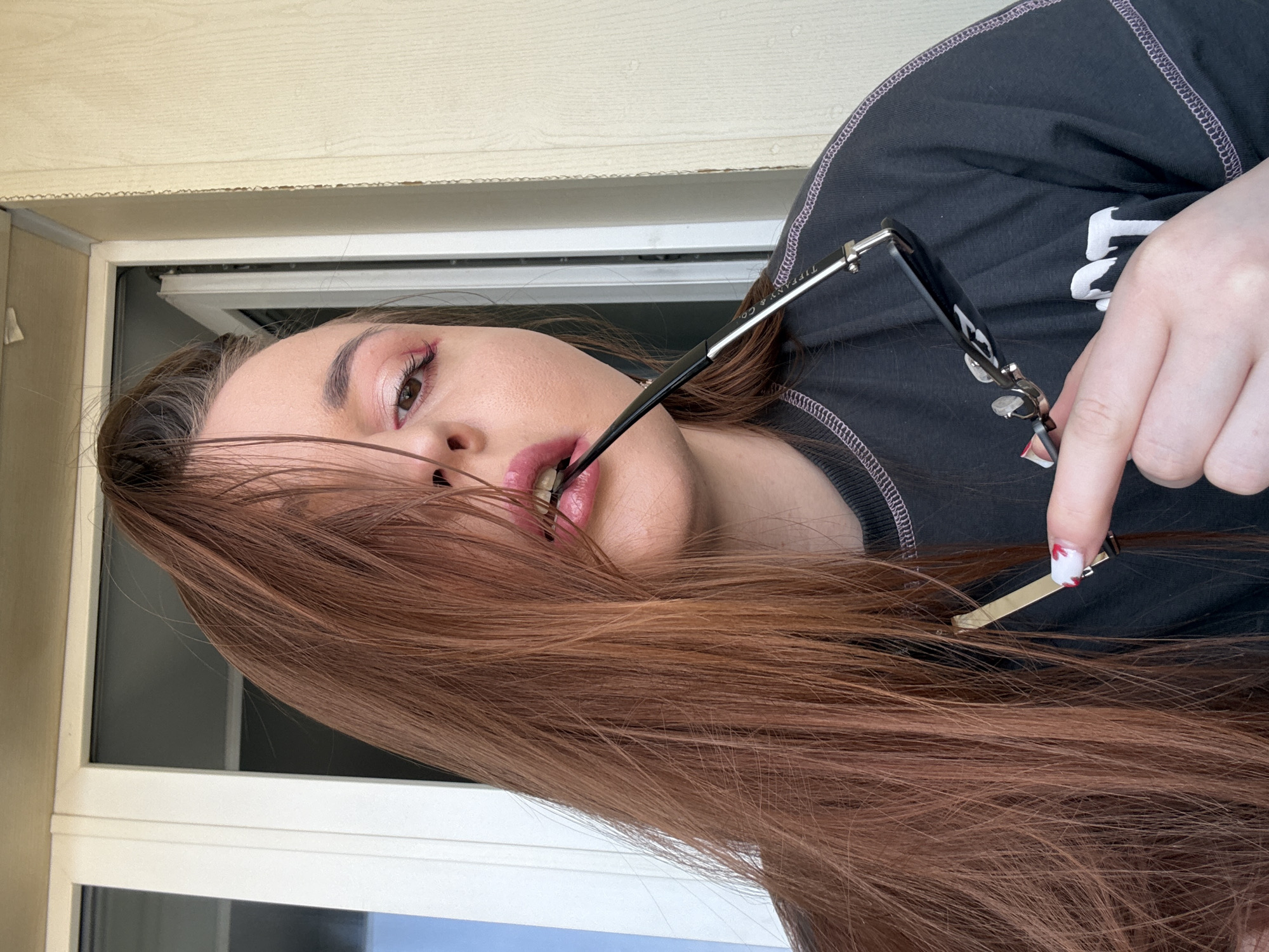A woman with long reddish-brown hair is positioned indoors near a window frame. She holds a pair of glasses in her mouth and hand, displaying manicured fingernails with a decorative red and white design. She is wearing a dark colored t-shirt with white lettering, posing against a window frame. The photo was not taken in Izhevsk, Russia.