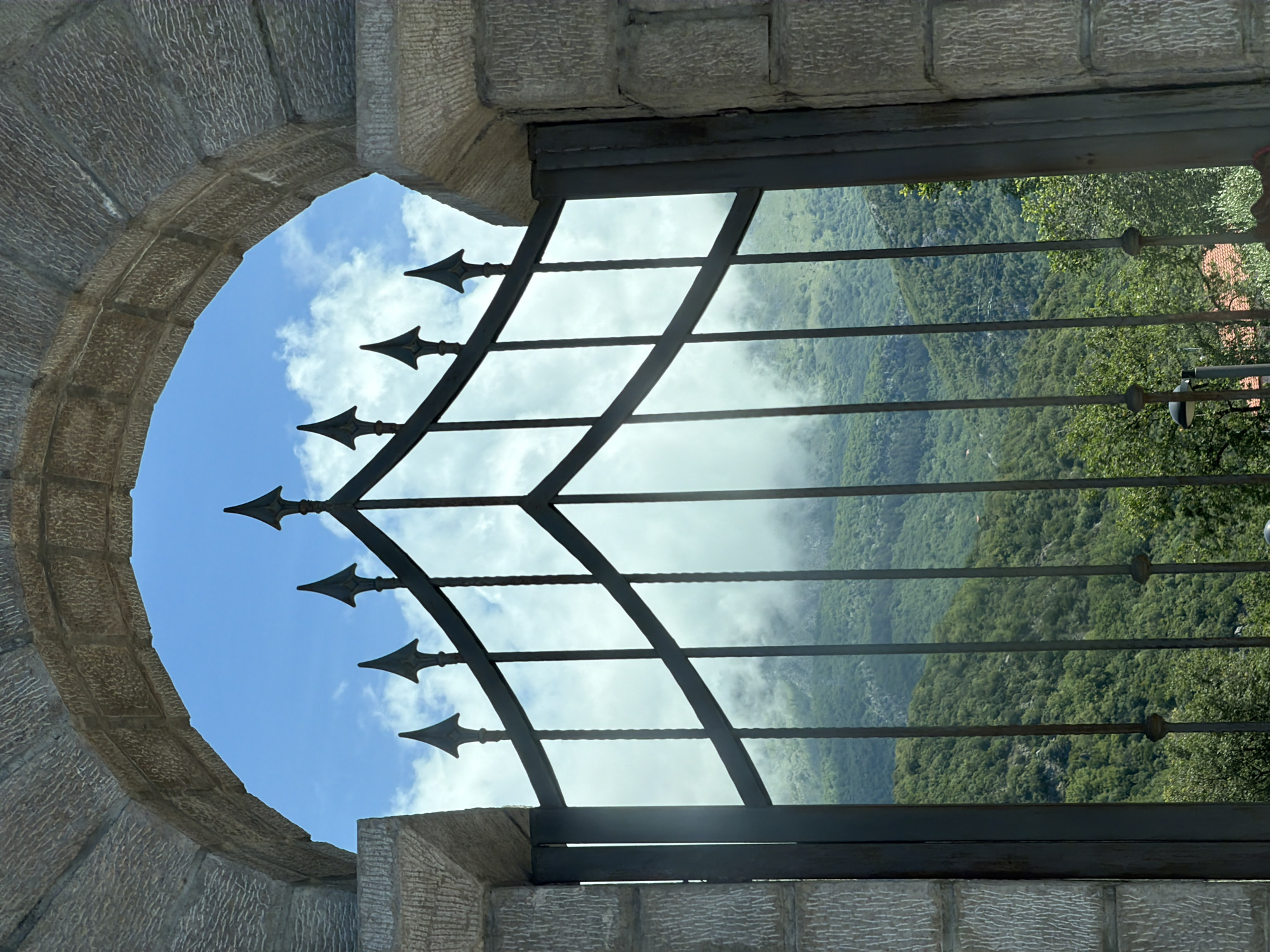 The image captures a scenic view through a stone archway and a metal gate, set in Kubasi, Montenegro, during daylight hours.
In the foreground, a robust stone archway, constructed from textured, light-colored blocks, frames the scene. The inner surface of the arch shows a rough, chiseled finish. Positioned within this archway is a dark metal gate, possibly wrought iron. The gate features a grid-like pattern of horizontal and diagonal bars, punctuated by multiple decorative, arrow-shaped finials pointing upwards, lending it a historical or protective appearance.
Beyond the gate, a vast natural landscape is visible. The sky above is bright blue with scattered white, puffy clouds. The dominant feature of the background is a series of rolling mountains densely covered in lush green forest and vegetation. Patches of white mist or low-hanging clouds partially obscure sections of the distant mountainous terrain and valleys, suggesting a high elevation or specific atmospheric conditions.
The overall scene conveys a sense of looking out from a secure or ancient structure onto a wild, expansive, and somewhat mysterious natural environment. There is no visible text in the image.