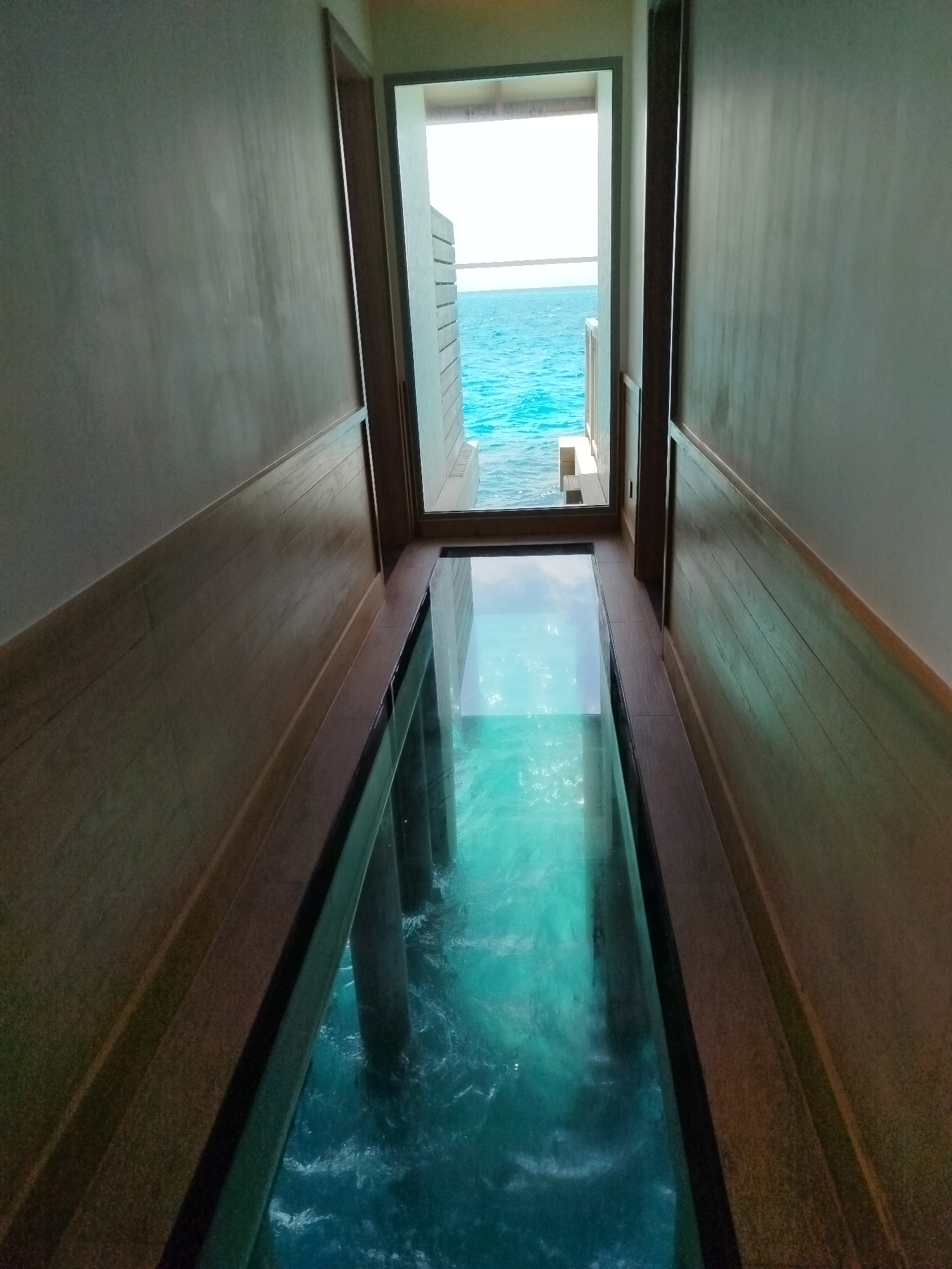 The image depicts a walkway with a glass floor, revealing the turquoise ocean water and supporting pillars below. The walkway is bordered by wooden paneling on the walls and a wooden railing. At the end of the walkway, a doorway opens to an external view of the bright blue ocean and sky, with the distinctive architecture of what appears to be a resort or overwater bungalow in the Maldives. The scene is well-lit, suggesting daytime. There are no people visible in the image. The location is identified as Kendhoo, Maldives.