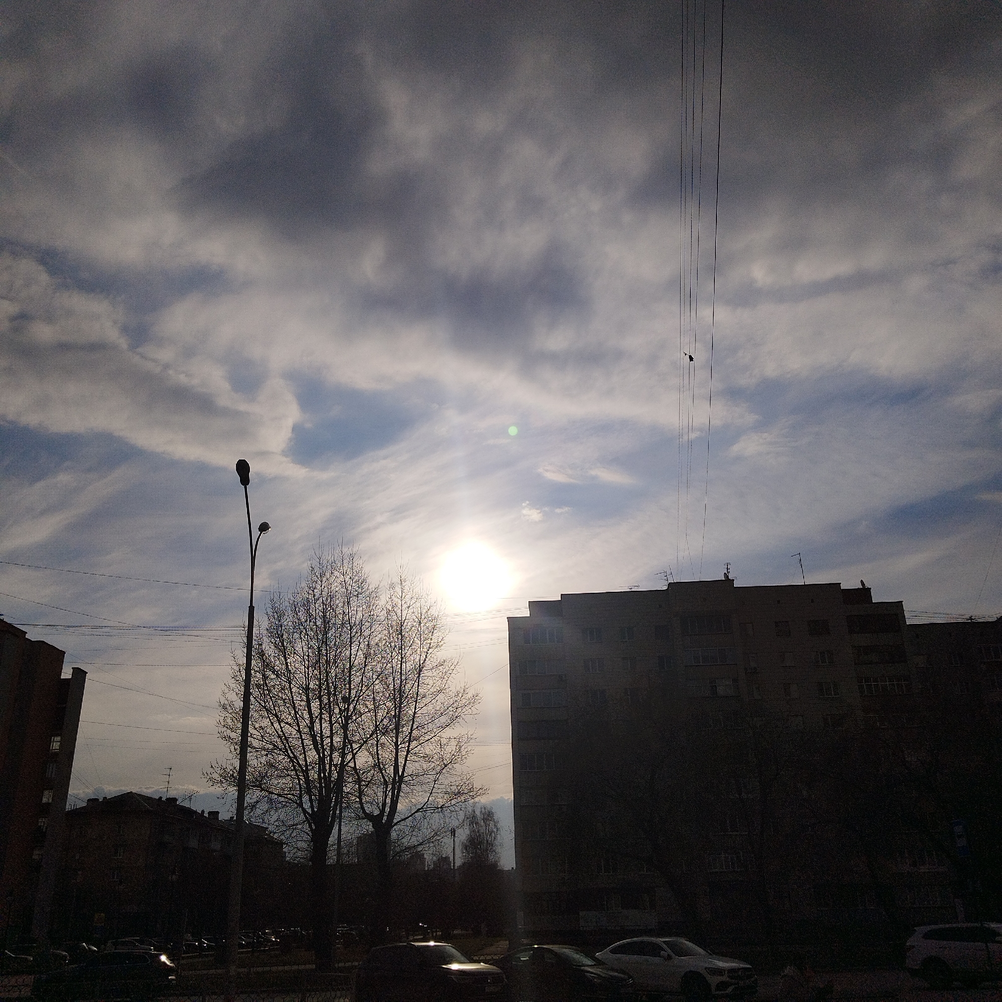 A wide-angle shot shows a city street in Yekaterinburg, Russia, at an overcast, bright time of day. The sun shines through a layer of white and gray clouds. In the midground, bare trees stand to the left of an apartment building. A lamppost is positioned near the trees on the left. Power lines run across the top right portion of the sky. In the foreground, parked cars are visible. Buildings are silhouetted on both sides of the street.