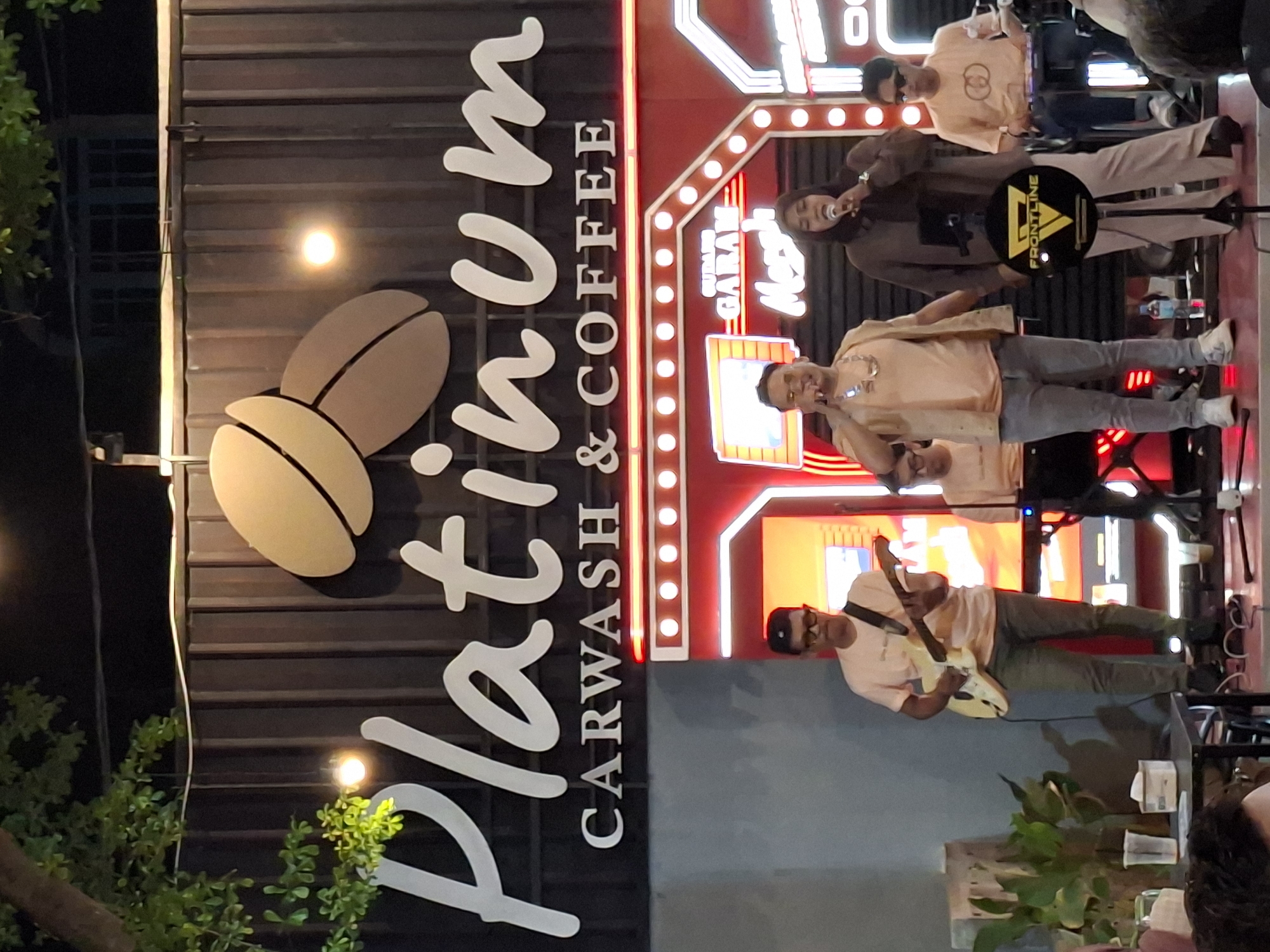 The image, taken at night in Lhokseumawe, Indonesia, captures a live musical performance at an outdoor or semi-outdoor venue. A prominent vertically oriented sign on the left displays "Platinum CARWASH & COFFEE" in large script and sans-serif fonts, accompanied by two stylized coffee bean graphics, indicating the establishment's services.
On a brightly lit stage to the right, several individuals are performing. In the foreground, a man in a light-colored shirt, dark pants, glasses, and a cap plays a light-colored electric guitar. To the right of the guitarist, another man in a light-colored shirt and grey pants sings into a microphone, while a woman in a dark top and possibly a head covering also sings into a microphone. Another man in a light shirt and sunglasses stands further back on the stage. A fifth person is partially visible below the male singer.
The stage is adorned with red strip lighting and features a yellow and black stand with the text "FRONTLINE". Other visible text on the stage backdrop includes "GARAM" and "Mangku", possibly indicating an event or brand. The performers appear engaged in their act, suggesting a lively atmosphere. The background consists of a dark corrugated metal wall, and some green foliage is visible in the lower left foreground. The artificial lighting clearly indicates the event is taking place after dark.