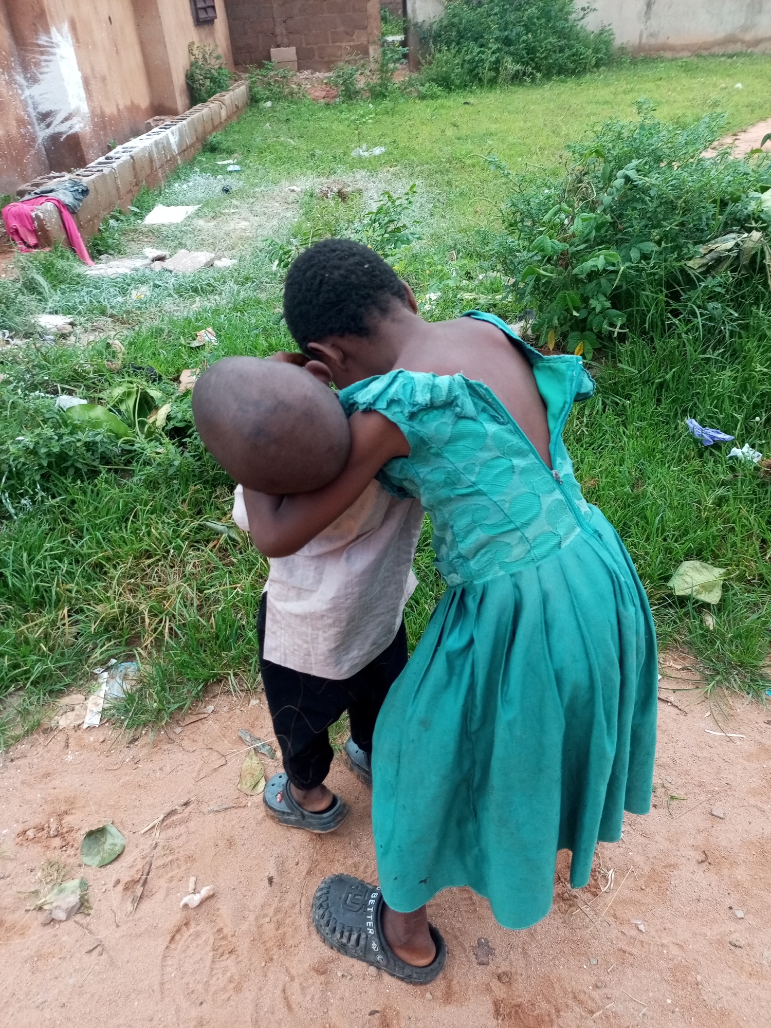 Two children, one wearing a teal dress and the other a light-colored shirt and dark pants, are interacting in an outdoor environment. The child in the teal dress is positioned in front and appears to be hugging or holding the other child. The child in the teal dress has a ruffled, off-the-shoulder design on their sleeves and a zipper down the back. They are also wearing dark-colored clogs. The child in the light-colored shirt has a visible bald head and is wearing dark pants and gray clogs. The background consists of unpaved, sandy ground with patches of grass and some scattered debris. In the background, there is a building with a light-brownish wall and a small window, as well as some overgrown green foliage. The scene is captured from a medium-high angle.