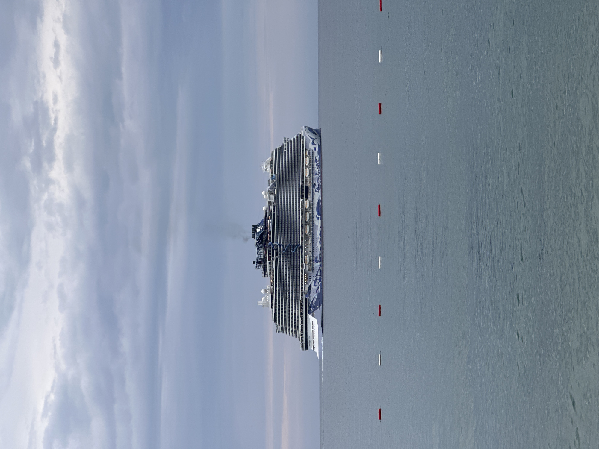 A horizontal shot reveals a cruise ship positioned in the sea off the coast of Bar, Montenegro. The sky is filled with clouds, and the sea is marked with intermittent red and white buoys.