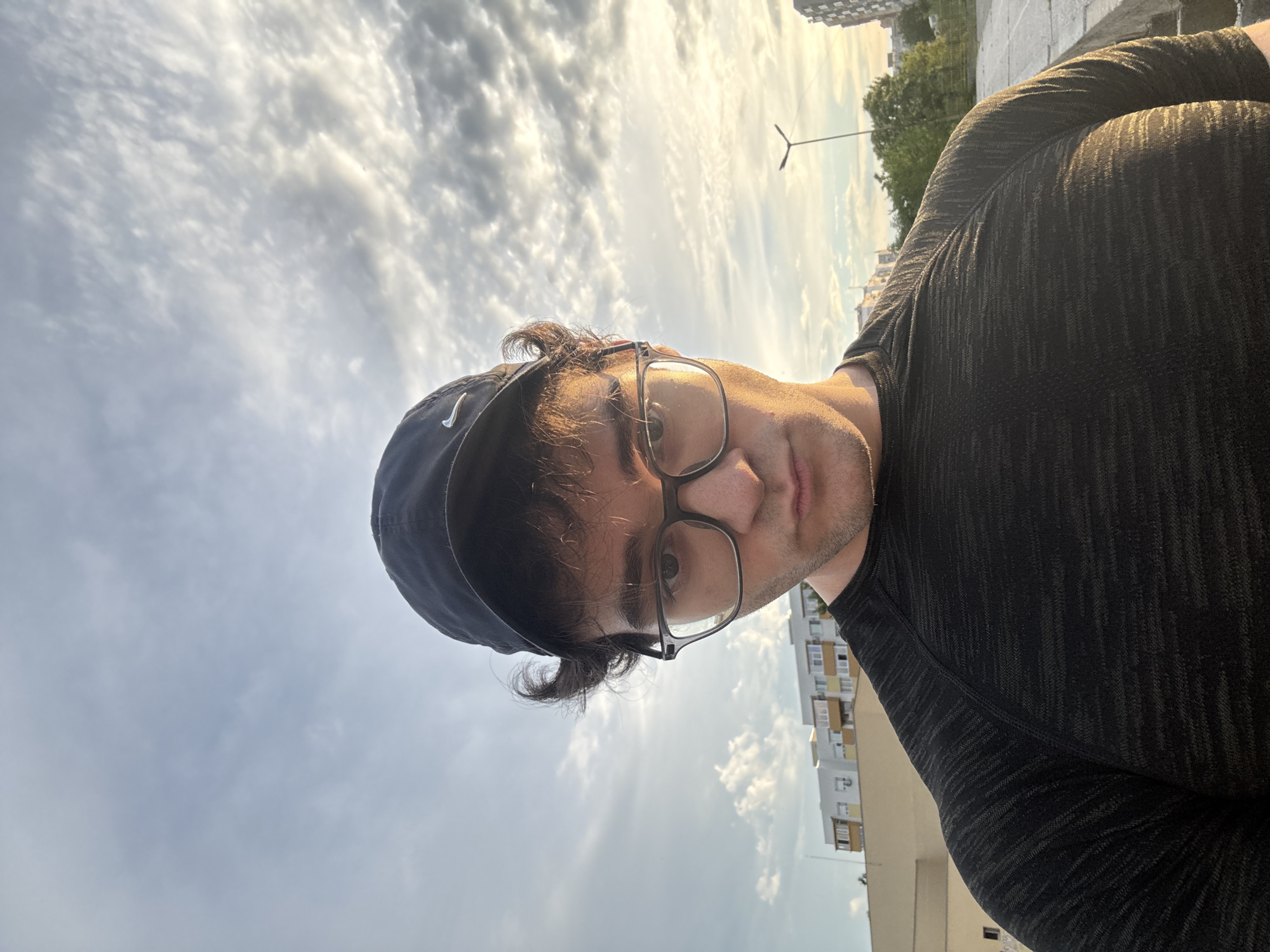 A medium-shot, eye-level selfie shows a young man wearing glasses and a black baseball cap with a Nike logo. He has curly brown hair and is looking directly at the camera with a neutral expression. He is wearing a dark, textured t-shirt. The background features a cloudy sky with the sun setting, casting a warm glow. In the middle ground, there are several apartment buildings and trees, suggesting an urban environment. The man is standing on what appears to be a concrete structure, possibly a rooftop or a walkway. The scene is outdoors in Blagoevgrad, Bulgaria, likely during the late afternoon or early evening.