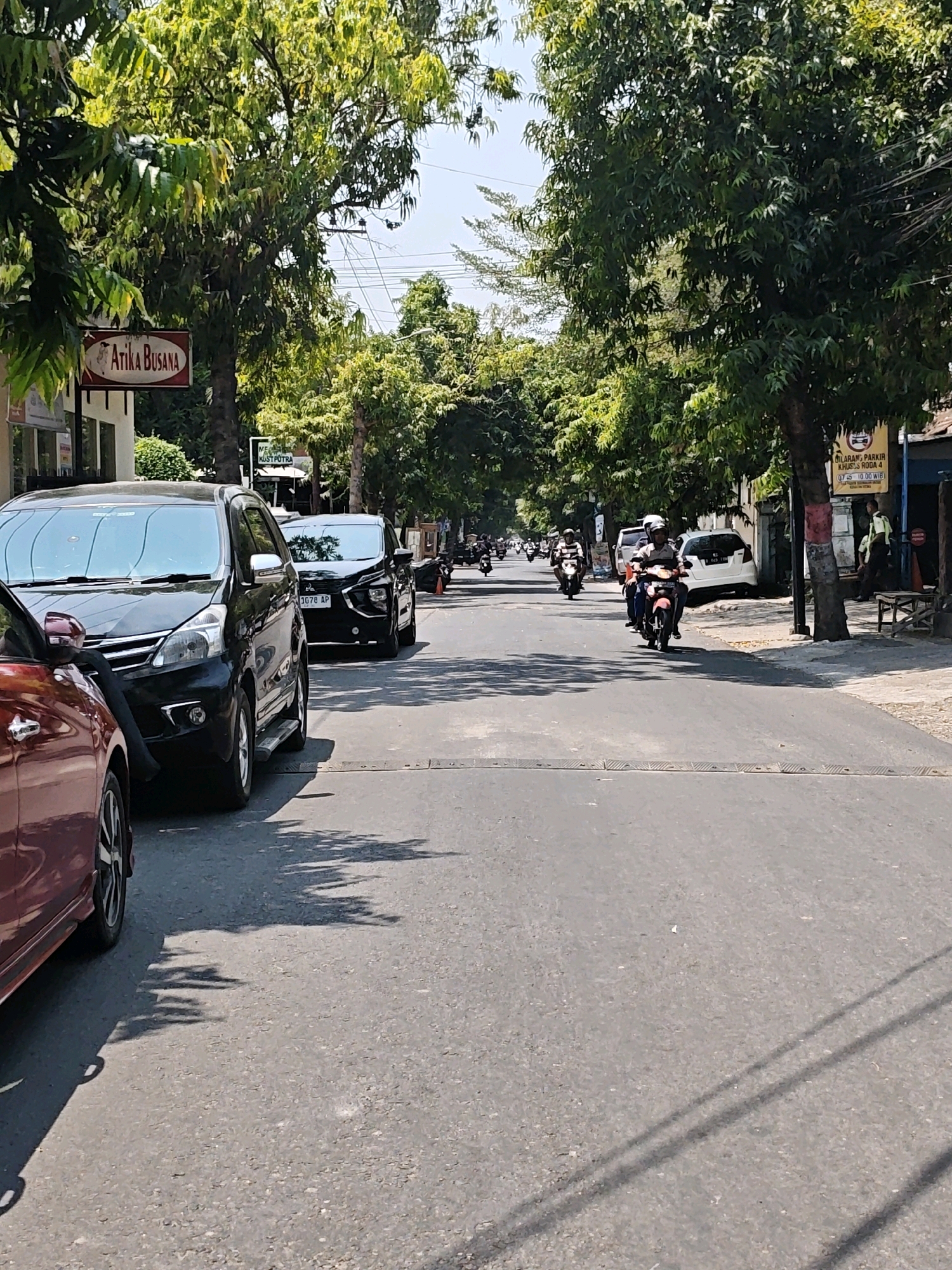 A street scene in Kediri City, Indonesia, features a two-lane asphalt road lined with trees on both sides. Several parked cars, including a black SUV on the left and another black car further down the road, are visible. A red car is parked on the far left. Multiple individuals are riding motorcycles, moving in the same direction as the parked cars. Buildings and shops are present along the street. The sky is clear and blue, indicating a sunny day. A sign with the text "Atika Busana" is prominently displayed on a building on the left. Another sign with "LARANGAN PARKIR" is visible on the right. The overall atmosphere suggests a typical day in a populated urban area.