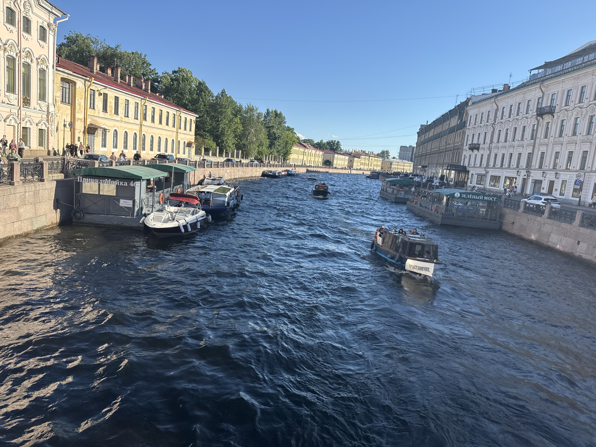 This is a photograph of the Griboyedov Canal in Saint Petersburg, Russia. The canal runs through the city center, lined with historic buildings. In the foreground, a waterway is filled with choppy blue water and several boats. Some boats are docked along the embankment, while others are in motion, navigating the canal. On the left bank, a long, light yellow building with many windows and red-tiled roofs stretches along the canal. Below the building, a stone embankment is visible, with a walkway and black metal railings. Several people are walking along this embankment. Further down the canal, more buildings line the right bank, one of which is a light-colored building with many windows and decorative architectural elements. The sky is clear and blue, indicating a sunny day. The scene captures a vibrant urban waterway with both historical architecture and contemporary river traffic.