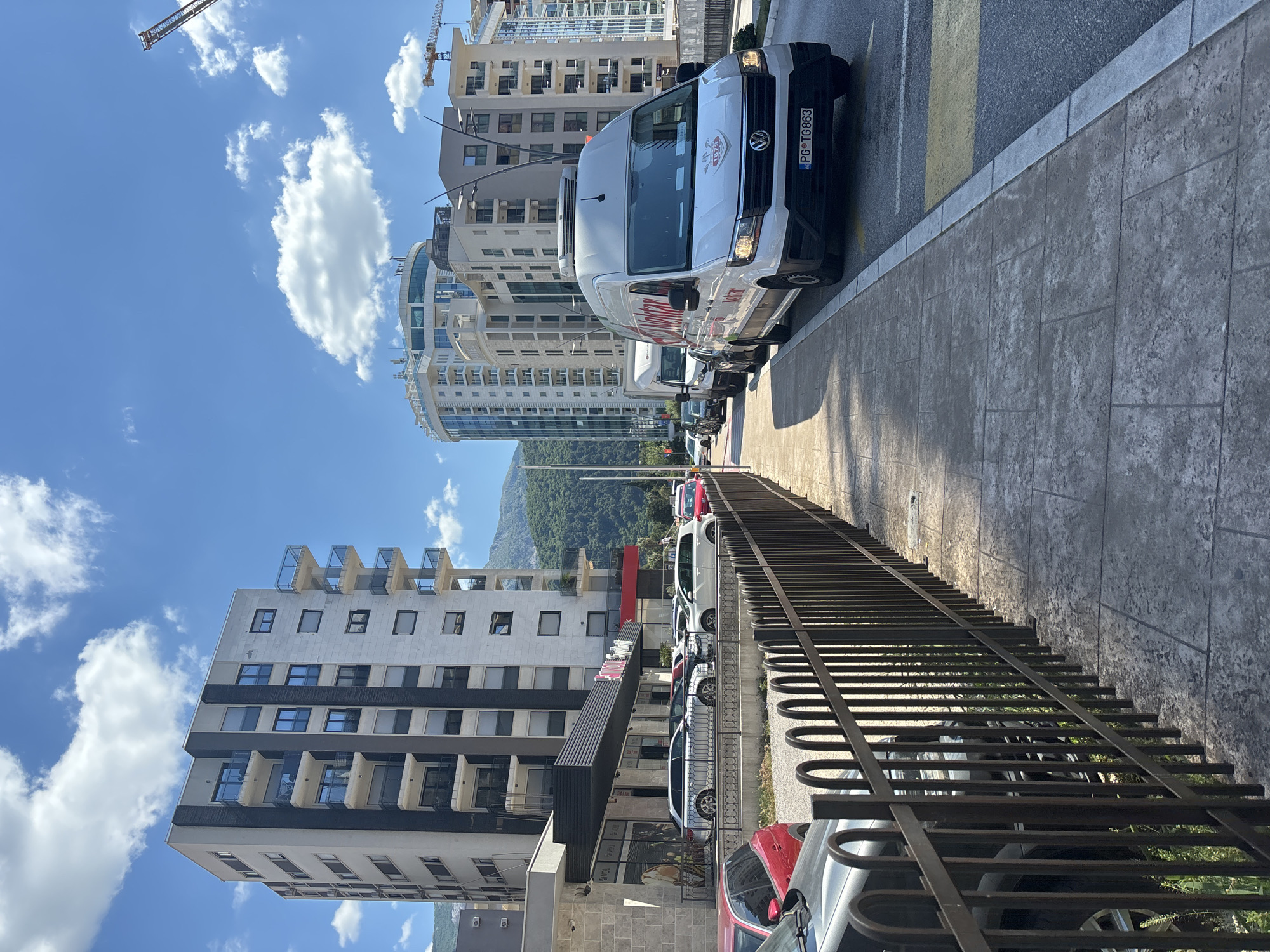 The image shows a street scene in Budva, Montenegro on a sunny day. A white Volkswagen van with the license plate "PG TG863" is parked on the side of the road. Several other cars are parked behind it. A black metal fence runs along the sidewalk, partially obscuring the parked cars. In the background are several tall buildings and a distant green mountain. The sky is blue with scattered white clouds. A construction crane is visible in the upper left corner of the frame.