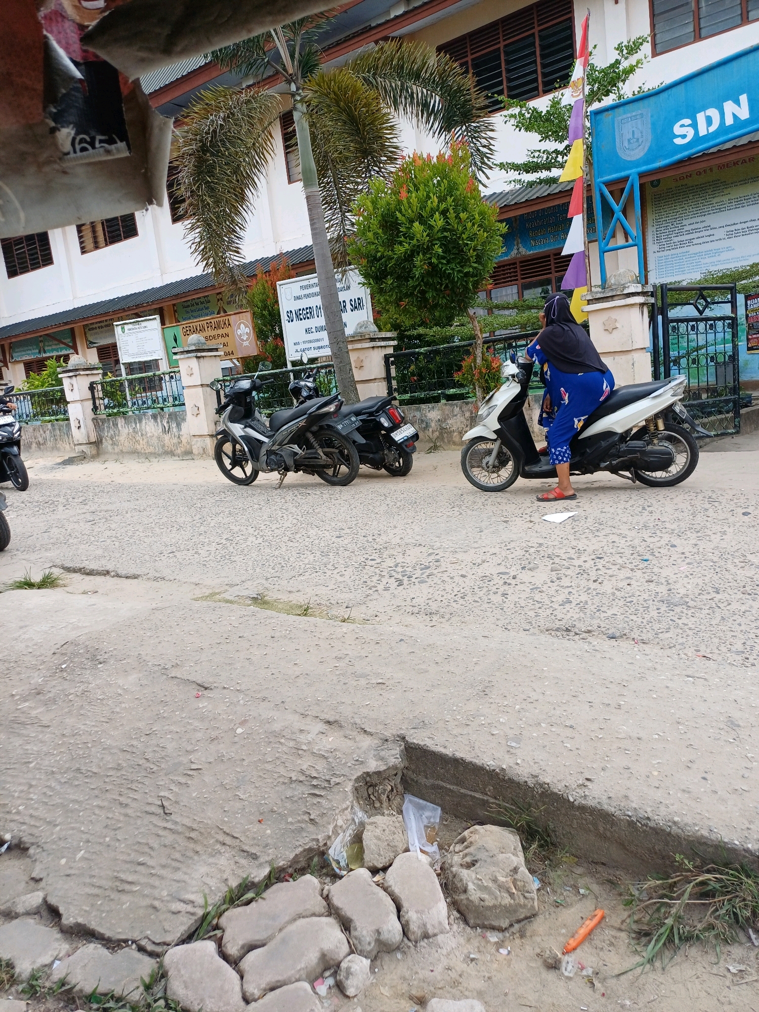 The image depicts a street scene in front of a school building, identified by signs as "SD NEGERI 01 MEKAR SARI" and "DINAS PENDIDIKAN KOTA DUMAI," indicating its location in Dumai City, Indonesia. A woman, wearing a blue patterned dress and a black headscarf, is seated on a white scooter. She is looking to her left, with her right foot on the ground. Another scooter, silver and black, is parked to her left, and a black scooter is parked to the left of that. In the background, palm trees and foliage are visible, along with a building that appears to be the school. The street is paved with asphalt, and there is a damaged section of concrete sidewalk in the foreground with some loose stones and debris. An orange object, possibly a cigarette lighter or candy wrapper, lies on the ground near the damaged sidewalk. The sky is not visible in the frame.