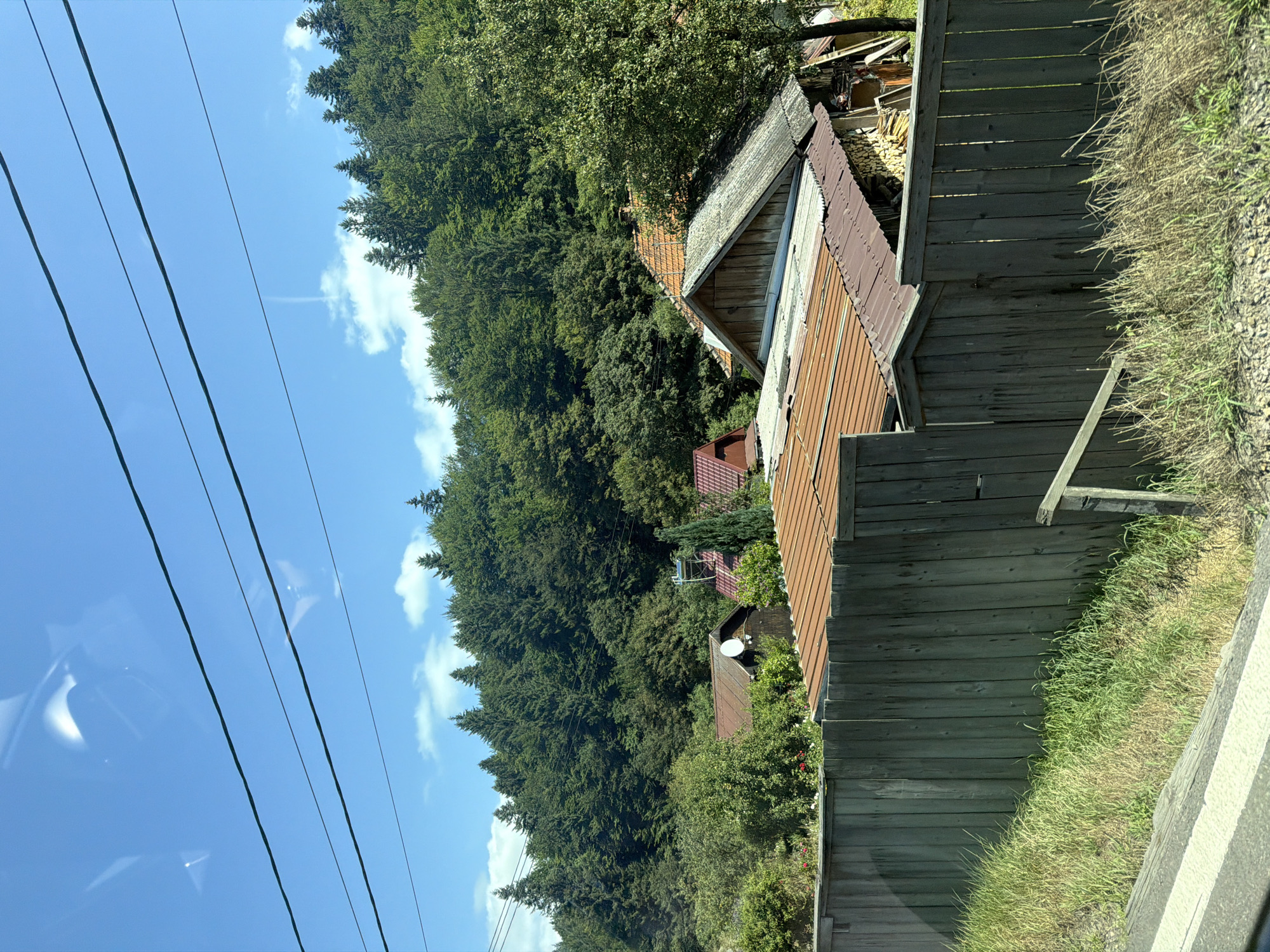 In Oituz, Romania, a wooden fence runs horizontally across the foreground, bordering an area with green grass and dry vegetation. Behind the fence, several rooftops are visible, some with orange tiles and others with corrugated metal. A small satellite dish is attached to the side of a dark wooden building with a red-tiled roof. Further back, a dense forest covers a hillside under a bright blue sky with scattered white clouds. Several power lines are strung across the upper portion of the frame, intersecting with the sky. The scene appears to be a rural or semi-rural residential area.
