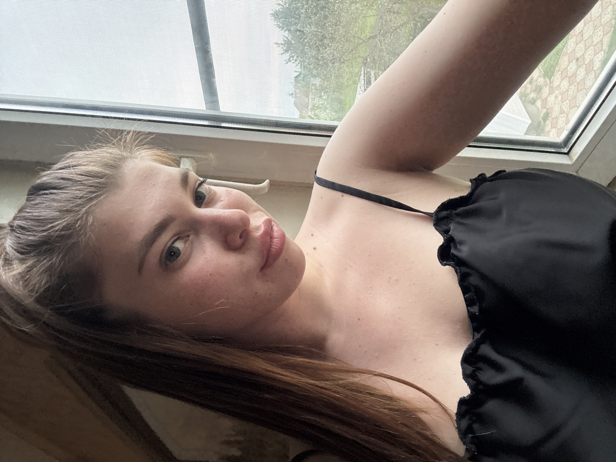A woman with light brown and blonde hair is posing for a selfie near a window. She is wearing a black top with a thin strap. The window has a screen and the view outside includes greenery. The location is not discernible from the image.