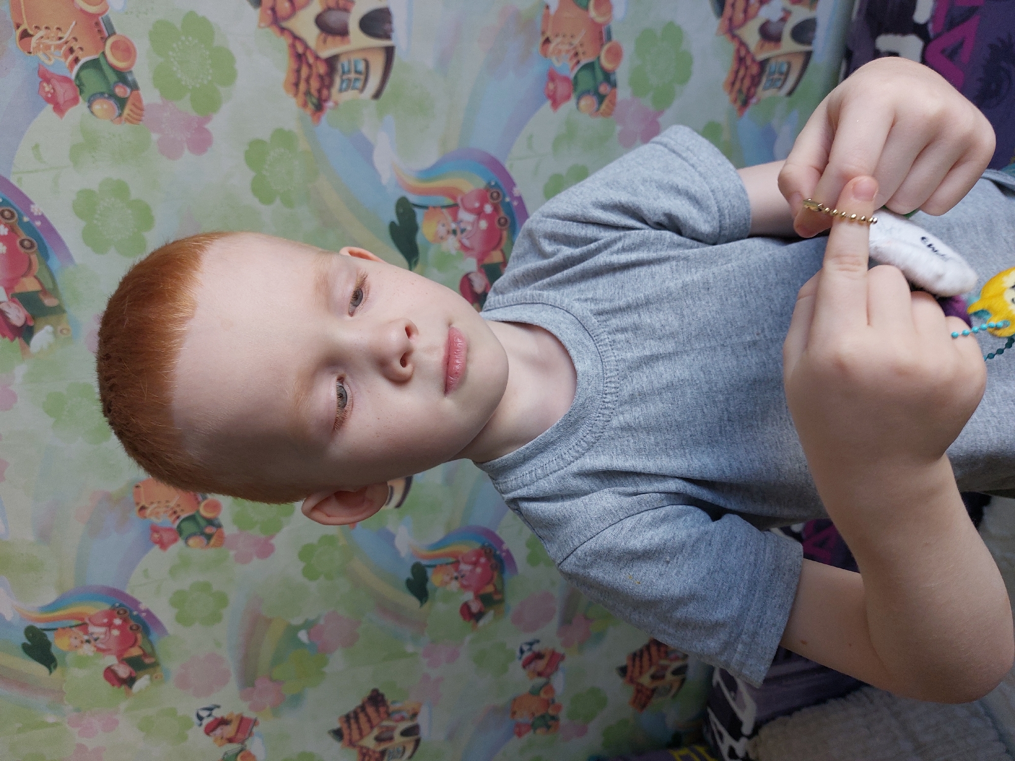 A young child with fair skin and red hair is lying down, wearing a gray short-sleeved shirt. They are holding a small, white, plush object attached to a gold chain. The background consists of wallpaper with a pattern of cartoon figures, flowers, and rainbows. Based on the location metadata, this scene is likely located in the city of Chaikovsky, Russia.