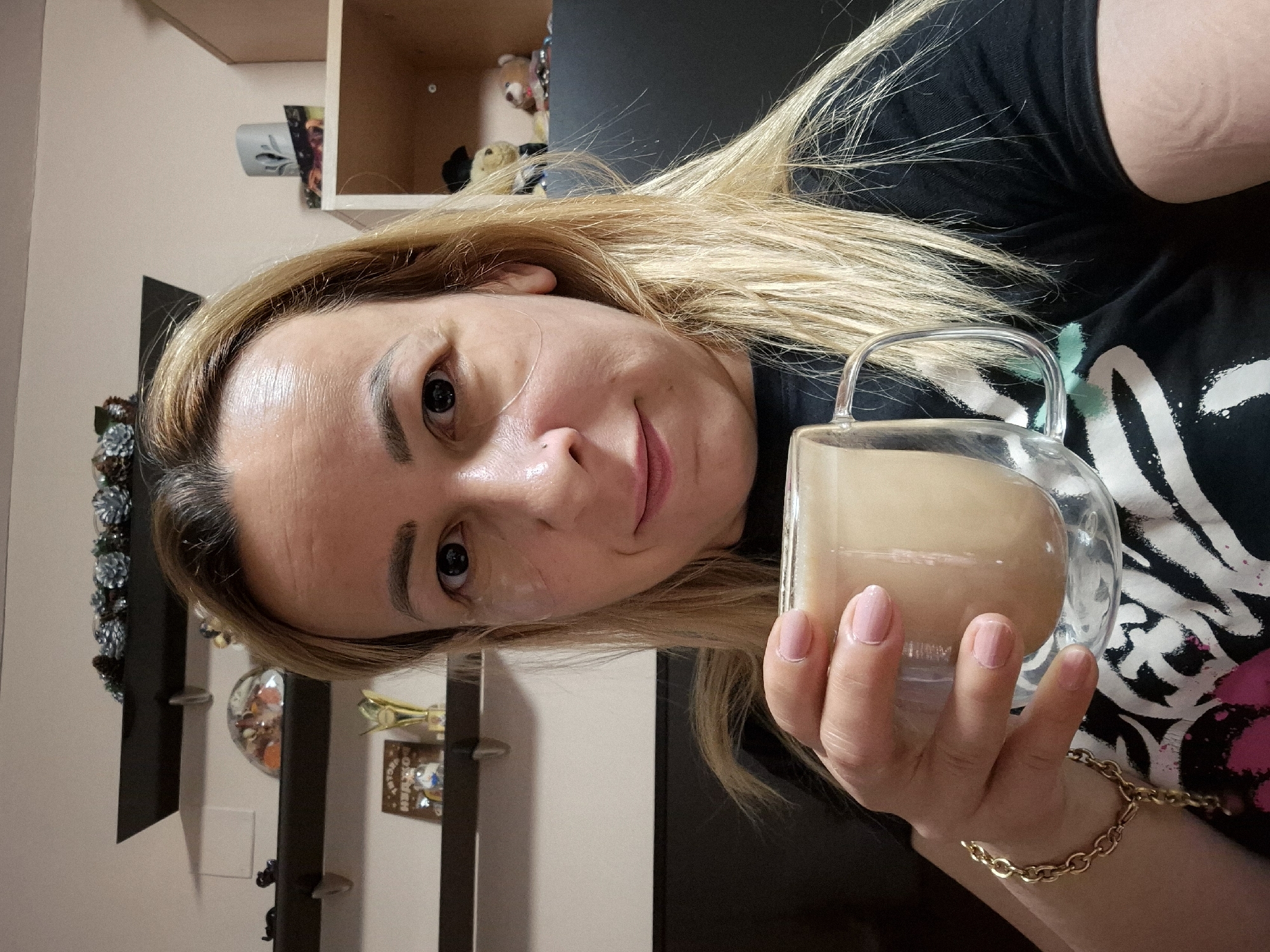 A woman with blonde hair and light skin is holding a clear glass mug filled with a light-colored beverage, possibly coffee or tea, in her right hand. She is looking towards the camera with a slight smile. She appears to be wearing under-eye patches and has a gold bracelet on her left wrist. She is wearing a black t-shirt with a white and pink design. The background shows shelves with decorative items and stuffed animals. The image appears to be taken indoors, possibly in a home.
