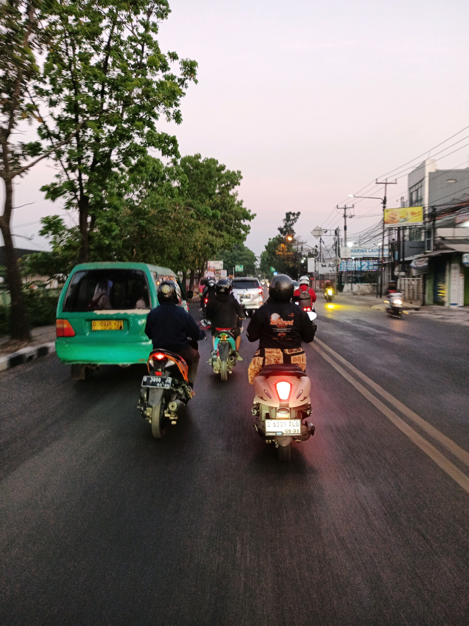 The image depicts a street scene in Bandung City, Indonesia, likely during the late afternoon or early evening, given the soft light and the presence of streetlights and illuminated signs. The perspective is from behind several motorcycles traveling on a paved road.
In the foreground, two motorcycles are prominently visible. The closest one, on the right, is a light-colored scooter with its brake light illuminated, indicating it is slowing down or stopped. The rider is wearing a black jacket with a graphic on the back and a helmet. Behind this scooter is another motorcycle, dark in color, also with its brake light on. Its rider is wearing a dark jacket and helmet. Further ahead and to the left, another motorcycle with a rider in a red jacket and helmet is visible.
To the left of the road, a bright green van is stopped or moving slowly, with its rear visible. The van has a license plate that reads "D 4928 ZS". Several people appear to be inside the van, though their features are indistinct.
The road itself is asphalt with yellow lane markings. Along the right side of the road, there are buildings with signs, including one for "HARINA LAUNDRY". Power lines and utility poles are visible overhead. Trees with green foliage line the left side of the road, suggesting a suburban or mixed urban-rural environment. The sky is a pale, overcast color, common in twilight or during cloudy weather. The overall impression is of moderate traffic flow in a bustling urban setting.