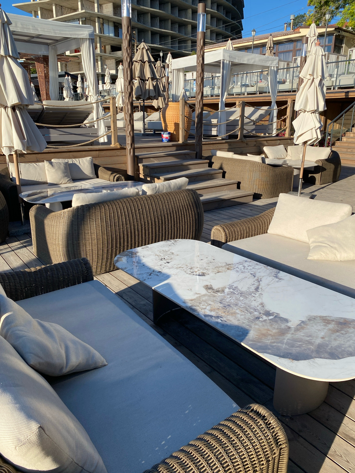 The image shows an outdoor seating area at a waterfront establishment in Odesa, Ukraine. The area features wicker furniture, including sofas and chairs, arranged around marble-topped tables. White umbrellas are set up to provide shade, and white canopies are visible in the background. Wooden decking and steps form the flooring. A building under construction is partially visible in the upper background. The sun is casting shadows across the scene, indicating it is daytime. There are no people visible in the image. The overall impression is of a leisure or dining space, likely part of a beach club or restaurant.