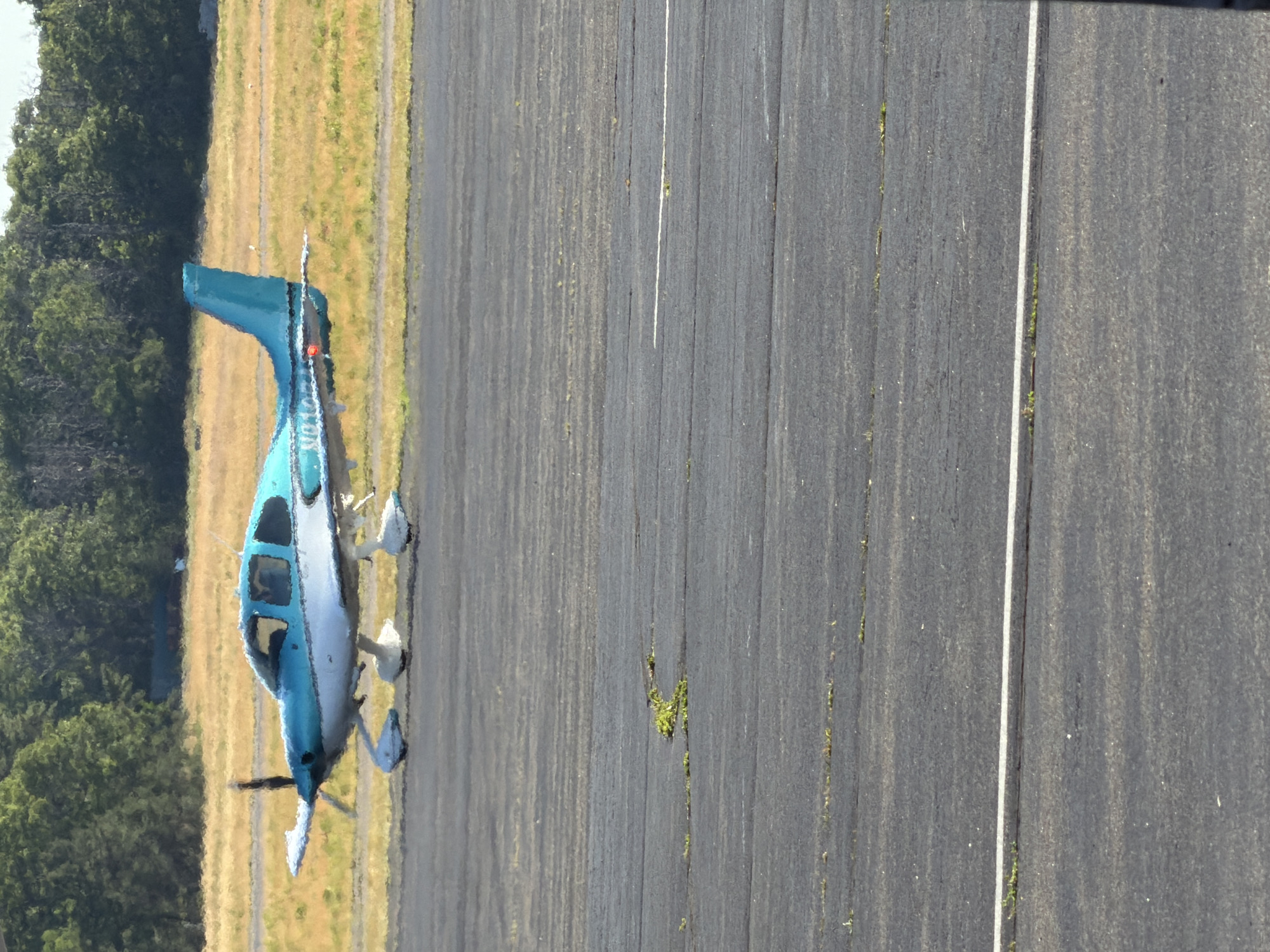 A small, light blue and white airplane is parked on an asphalt runway. The airplane has a single propeller at the front. The background consists of a grassy field with trees in the distance. The scene appears to be an airfield in the Town of East Hampton, United States of America.
