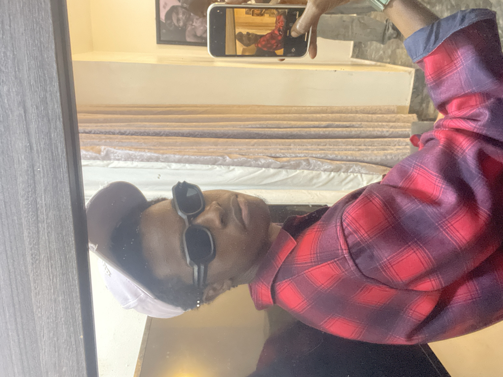 A person with dark skin, wearing a red and purple plaid shirt, black sunglasses, and a baseball cap with a white front, is visible in a mirror reflection. The person is holding a smartphone in their right hand, which is displaying a mirror image of the same person.
Behind the person is a stack of mattresses wrapped in plastic. Above the mattresses is a picture frame and some white, built-in shelves. The location of the scene is potentially in a mattress store or warehouse, possibly in Awka, Nigeria. The specific environment and objects suggest a retail or storage setting for mattresses.