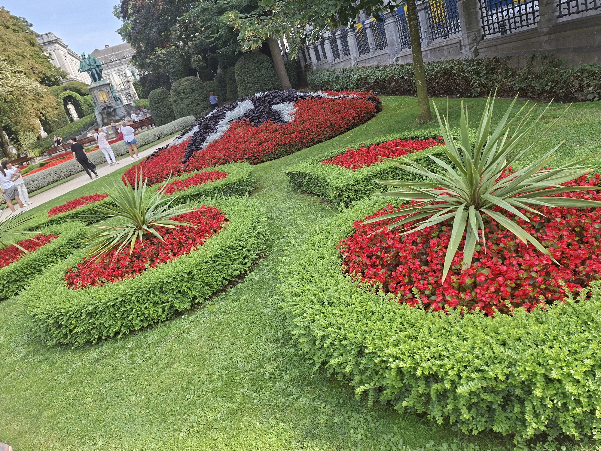 In this sunny outdoor shot, a meticulously maintained formal garden features a vibrant display of red flowers bordered by manicured green hedges. In the mid-ground, several people are visible strolling along a pathway, enjoying the scenery. Behind them, a prominent statue stands as a central focal point, with a grand building visible in the background. The park setting is lush with trees and grass. This scene appears to be a snapshot of the Jardin du Mont des Arts in Brussels, Belgium, a popular urban park known for its floral displays and city views.