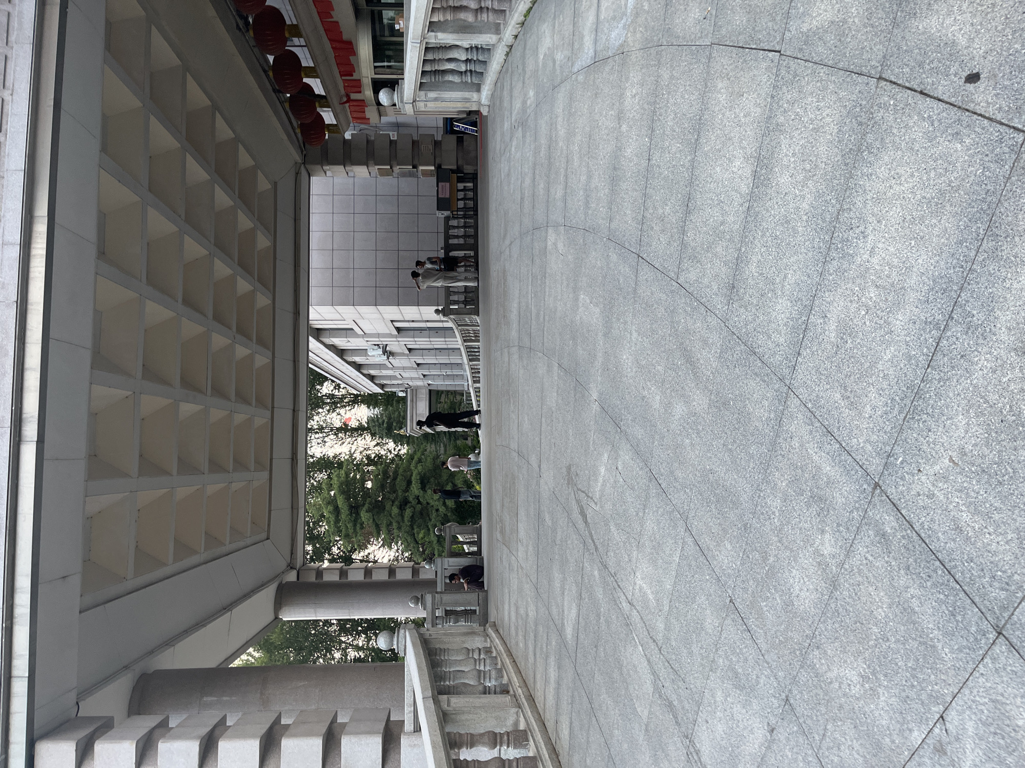 An outdoor, low-angle, full shot captures a wide, gently sloping stone ramp leading under a covered entryway of a building. The entryway is supported by large, square pillars and features a coffered ceiling. Red lanterns and red banner decorations hang from the right side of the covered area, suggesting a festive or official occasion. Several people are visible in the mid-ground. On the left, near a stone balustrade, a person sits on the ground. Further back, a group of individuals stands and converses. The building appears to be modern with a tiled exterior. Trees line the background behind the building. The overall scene suggests a public space in Changchun City, China, possibly a government building or a cultural institution, during daytime hours.