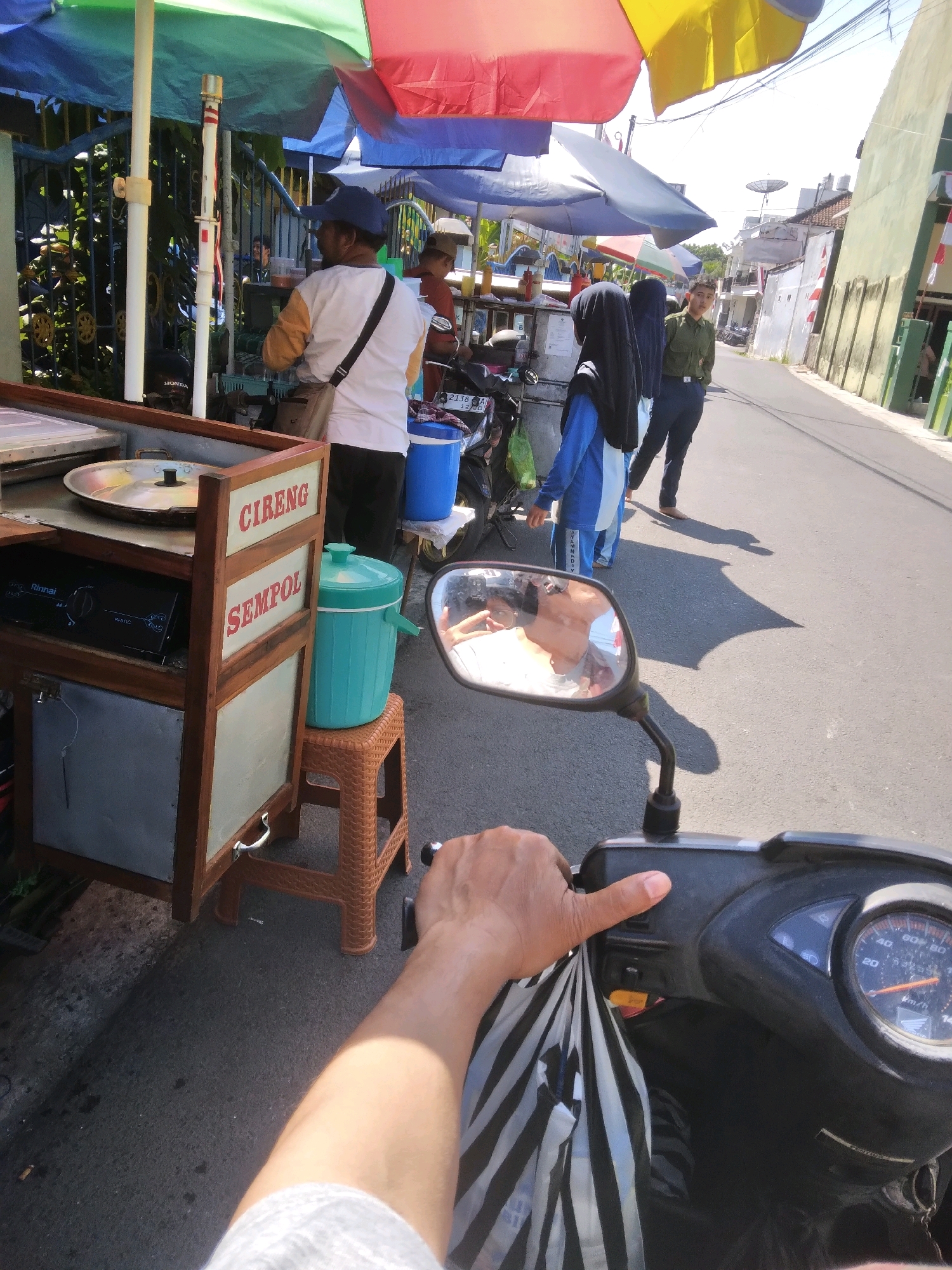 The image captures a street scene in Purwokerto, Indonesia, likely during daylight hours given the bright sunlight and clear sky. In the foreground, the perspective is from the rider of a scooter, with their hand visible on the handlebars and holding a black and white striped bag. The scooter's dashboard and speedometer are also visible. To the left, a mobile food stall is set up, characterized by a wooden cart displaying "CIRENG SEMPOL" signage and a gas stove. Behind the food stall, a person wearing a blue headscarf and a blue and white outfit is standing, facing away from the camera. Another person in a black headscarf and blue outfit is partially visible behind them. Further down the street, a person in a dark green uniform and dark pants is standing, looking towards the left. The street is paved, and buildings line the right side of the road. Multiple umbrellas, some colorful, are set up over the food stall area, providing shade. A bright blue water dispenser is placed on a woven stool next to the food cart. In the reflection of the scooter's side mirror, a person is visible, appearing to be taking the photograph.