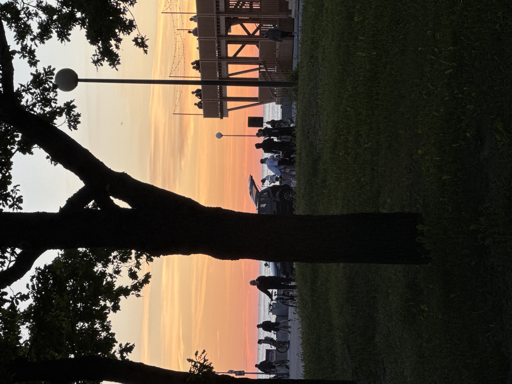 The image depicts a sunset scene near the coast in Tallinn, Estonia, likely along a promenade or park area. In the foreground, silhouetted trees cast shadows across a grassy area. A paved pathway runs parallel to the water, where several individuals are present. Some people are standing with bicycles, while others are engaged in conversations or observing the surroundings. A dark-colored car with its trunk open is parked near the pathway, with a group of people gathered around it. Further in the background, a multi-level wooden structure, possibly a pavilion or viewing platform, is visible, with people standing on its upper level. String lights are visible above this structure, adding to the evening ambiance. The sky is filled with the warm hues of a sunset, with orange and yellow tones dominating the horizon. The sea is calm and reflects the colors of the sky.