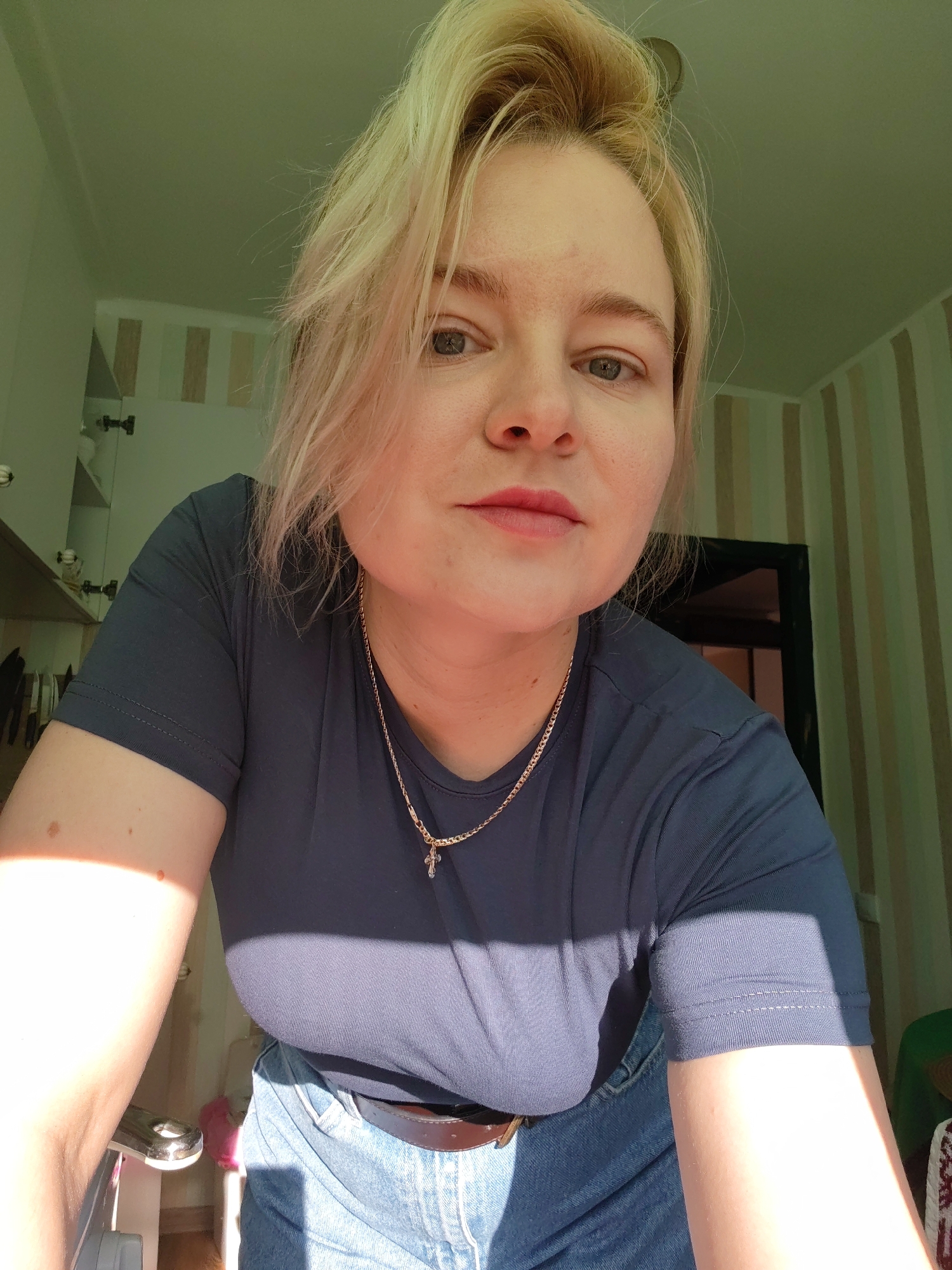 An indoor, medium-close shot shows a fair-skinned woman with blonde hair, wearing a dark blue t-shirt and blue jeans. She is looking at the camera, with her head tilted slightly forward. Her lips are slightly parted, and she has a light pink lipstick on. A gold chain with a small cross pendant rests on her chest. Her right arm is visible, with sunlight casting a bright glare on it, highlighting a few moles. In the background, vertical striped wallpaper is visible on the right, and a doorway is partially obscured by shadow. On the left, a shelf unit with some items can be seen. The lighting suggests a sunny day. The scene appears to be a domestic interior.