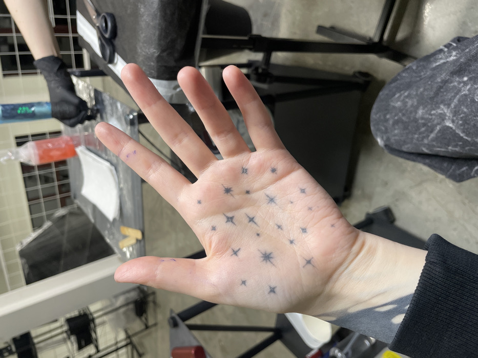 A close-up image displays a pale-skinned left hand, palm facing the viewer, featuring approximately 20 small, dark grey, four-pointed star tattoos scattered across the palm. Small purple marks are visible on the fingers. A larger, dark grey, abstract tattoo design extends onto the inner wrist and forearm, partially covered by a black long-sleeved garment. Patterned grey fabric, likely pants, is visible below the wrist.
The background depicts an indoor environment consistent with a tattoo studio. To the right, a black upholstered tattoo chair or bed is visible, with black scissors resting on its surface. Behind the hand, a grey workstation table is covered with a clear protective sheet. Objects on the table include folded white paper towels, a clear bottle containing red liquid, a blue spray bottle, and small wooden sticks. In the upper right background, a person's arm, gloved in black, is partially visible, holding a black tattoo machine over a surface. Metal shelving units and a window with a grid pattern are present in the blurred background. The floor is light grey, resembling concrete.
The specific location within Moscow, Russia, cannot be determined from the visual information provided in the image.