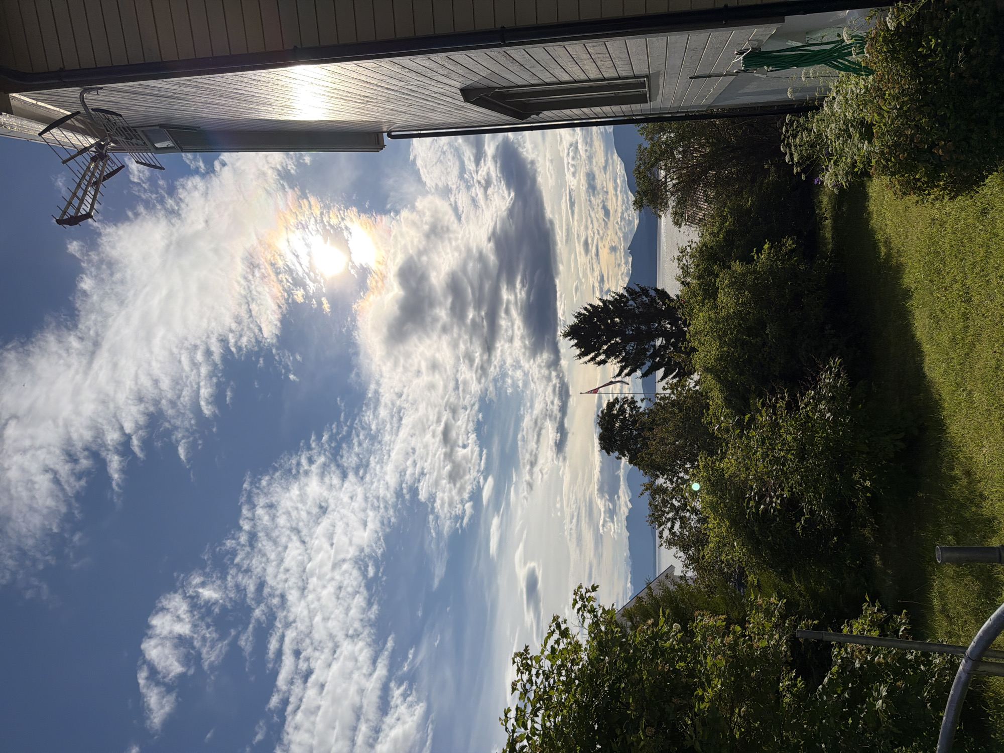 A bright, partly cloudy sky with the sun shining through, creating a rainbow effect, is visible above a body of water and distant mountains. In the foreground, the side of a light-colored house with horizontal siding is seen, along with a television antenna mounted on the wall. Green trees and bushes are in front of the house, and a patch of green grass is in the lower right quadrant of the image. A green garden hose is coiled on the side of the house. The scene appears to be a residential area in Harstad, Norway.