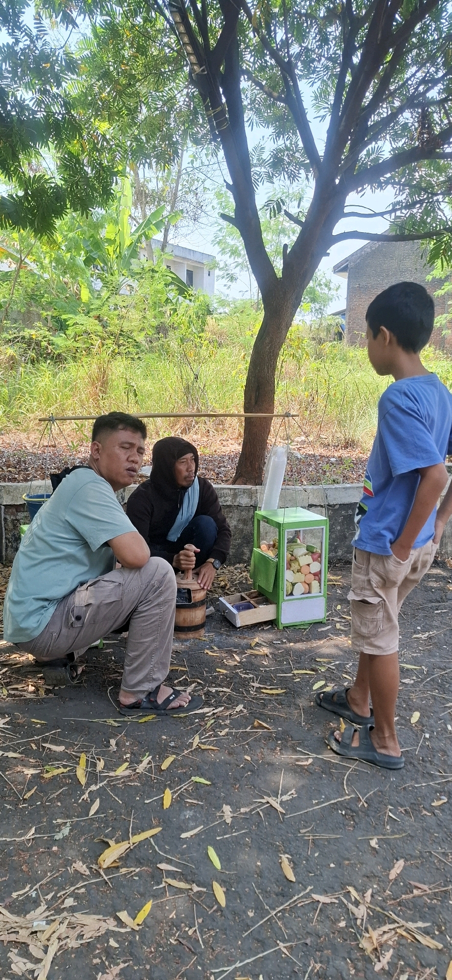 In Cilaku, Indonesia, three individuals are present in an outdoor setting. A man in a light blue t-shirt and beige cargo pants is squatting on the ground. Next to him, a person wearing a dark hooded jacket and a light blue scarf is also crouching and appears to be using a wooden mortar and pestle. In front of them, a boy in a blue t-shirt and beige shorts stands facing away from the camera, looking towards the other two individuals and a small green cart. This cart, likely a food or drink stand, contains cut fruits. The ground is a dark asphalt surface covered with fallen leaves and twigs. Behind the individuals, there is a low concrete wall, followed by overgrown green vegetation and trees. In the background, buildings are visible, suggesting a semi-urban or rural environment. The scene is illuminated by natural sunlight, casting shadows.