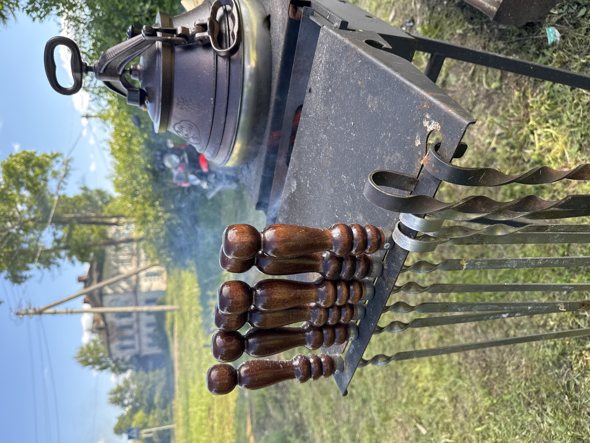 The image, rotated vertically, captures an outdoor cooking scene in Луптюг, Russia, during what appears to be a bright, clear daytime. The primary subjects are a large, dark metallic pot, resembling a kazan or Dutch oven, resting on a black metal brazier or grill stand. Smoke billows gently from beneath the pot, indicating an active fire heating its contents. The pot has a sturdy handle and lid, with some unreadable embossed patterns or text on its side.
Arranged on the side of the brazier are several metal skewers, featuring distinctive twisted shafts and dark, polished wooden handles. These skewers are typically used for grilling meat, such as shashlik.
The background reveals a rural or semi-rural setting, with vibrant green grass covering the ground and lush trees providing foliage. In the distant left, a traditional-style building, possibly an old house or estate, with visible windows and roof structures, is partially visible. A utility pole with power lines is also present. The clear sky with some clouds suggests pleasant weather.
No individuals are visible in the frame. The scene portrays an inviting outdoor cooking activity, likely preparing a meal such as a stew or soup in the pot, alongside grilled items using the skewers.