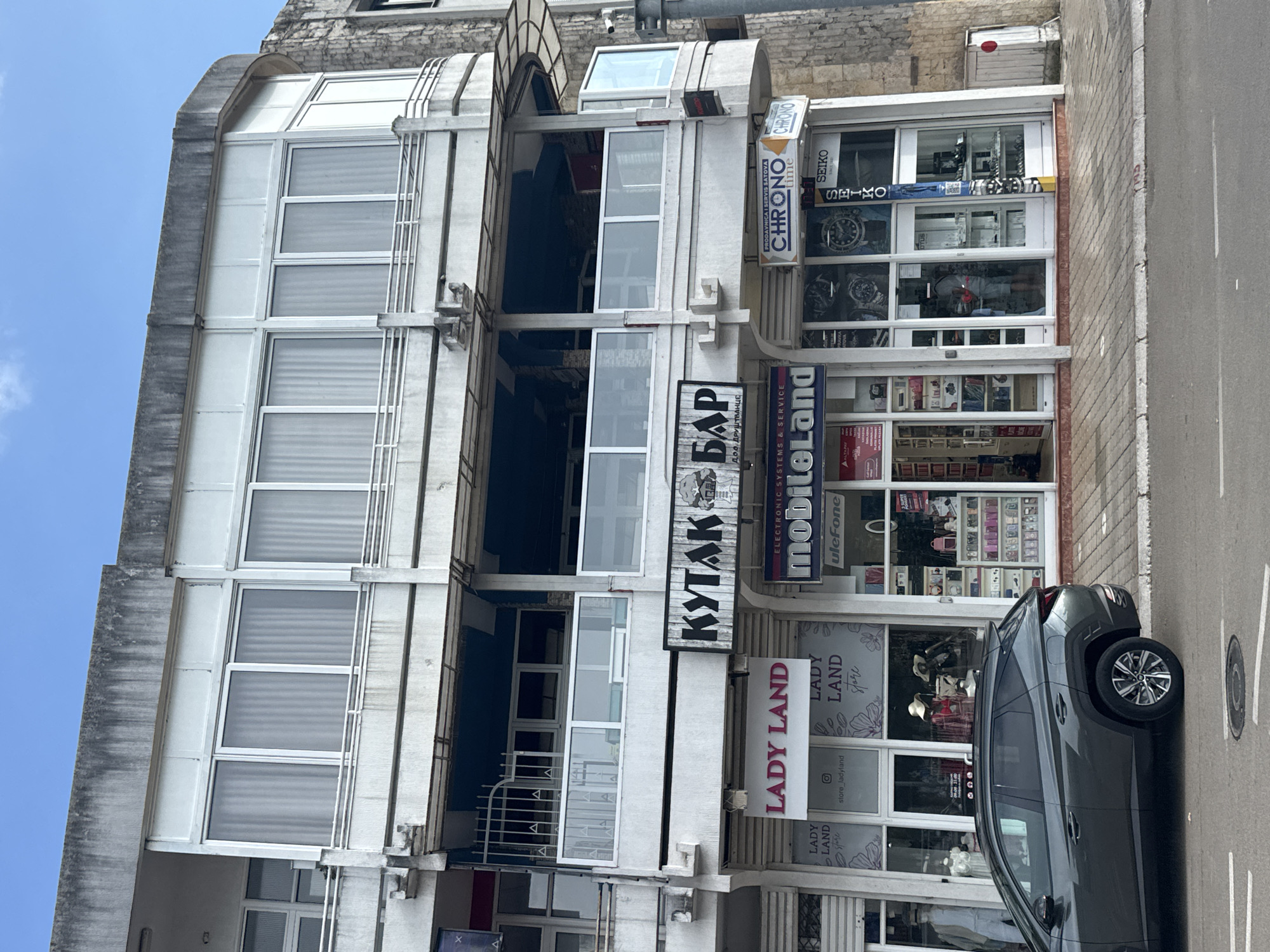 The image depicts a street view of a multi-story building with shops on the ground floor. The building appears to be located in Šušanj, Montenegro, as suggested by the shop signs. In the foreground, a dark gray SUV is parked on the street. The ground floor features several storefronts. One prominent sign reads "LADY LAND," with another "LADY LAND store" below it, displaying mannequins and merchandise within the shop. Adjacent to it is a shop with a sign that says "KYTAK БАР" (KYTAK BAR). Further to the right, there is a shop with the sign "CHRONO time" and "Seiko," showcasing watches. Another shop labeled "mobiloLand" with "uleFone" branding is also visible. The shops have large glass windows, some displaying products and mannequins. The architecture of the building is functional, with balconies and multiple levels of windows. The street is paved with cobblestones, and there are streetlights and various signs visible. The sky is clear and blue, indicating daytime.