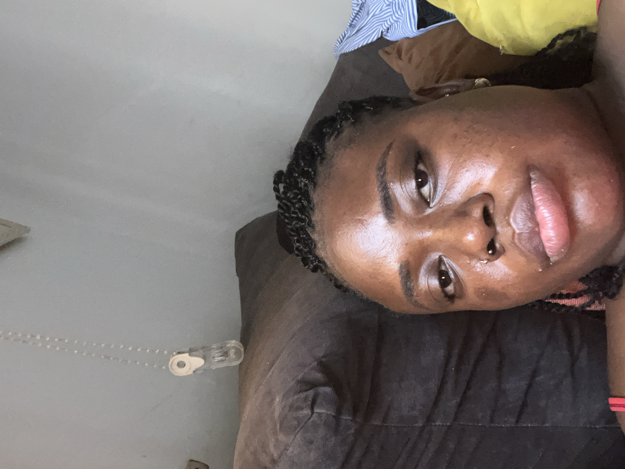 A close-up, overhead, indoor shot shows a Black person lying down on a dark brown sofa. The person's head and shoulders are visible. They have dark braided hair, a nose ring, and are wearing a red top. The background is a light-colored wall with a string of small, clear beads hanging down, attached to a white clip. The scene is likely a residential setting in Abuja, Nigeria.