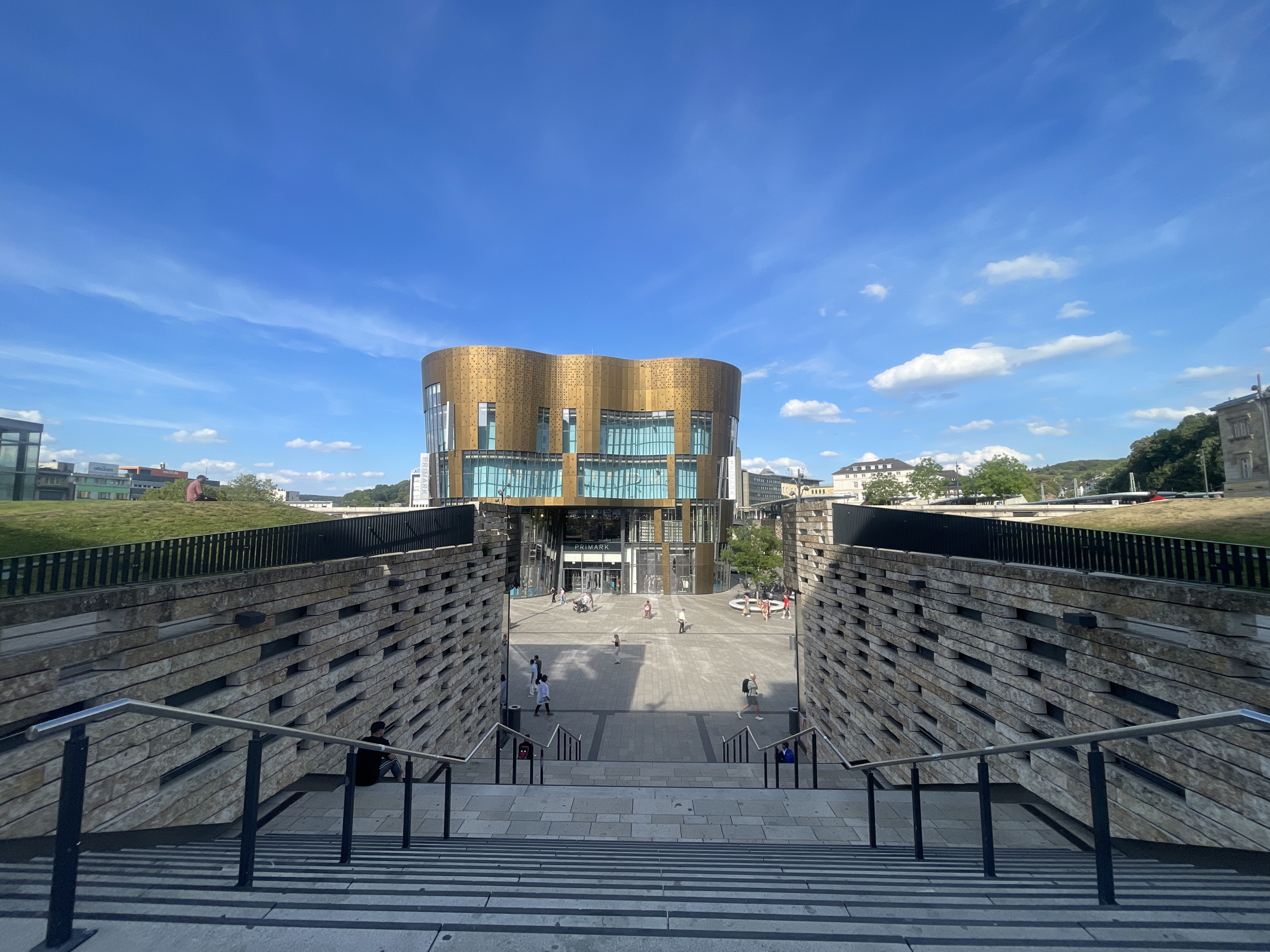 This is a ground-level, wide-angle shot of the Willy Brandt Square in Wuppertal, Germany. The square features a modern, curved building with a gold-colored facade and large glass windows, identified as "Primark" by signage above its entrance. The square is paved and has a fountain in the center. People are scattered throughout the square, some walking, others standing or sitting. In the foreground, a wide stone staircase with metal handrails leads down into the square. To the left and right of the staircase, there are sloped walls constructed of stacked stone blocks. The sky above is blue with scattered white clouds. Green grass and trees are visible in the background on the left and right.