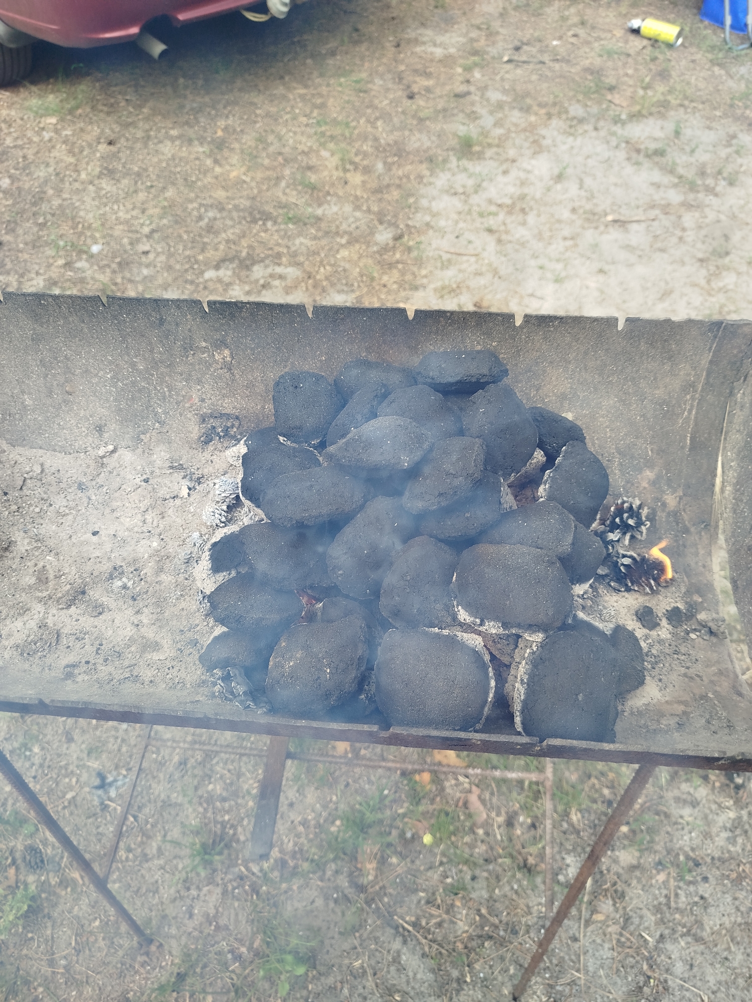 A pile of charcoal briquettes sits in a metal barbecue grill outdoors. Some of the briquettes are starting to glow red, and a small amount of smoke is visible. The grill is on a metal stand, and the ground around it is covered with dirt and some dry grass. A portion of a dark red car is visible in the upper left corner of the image.