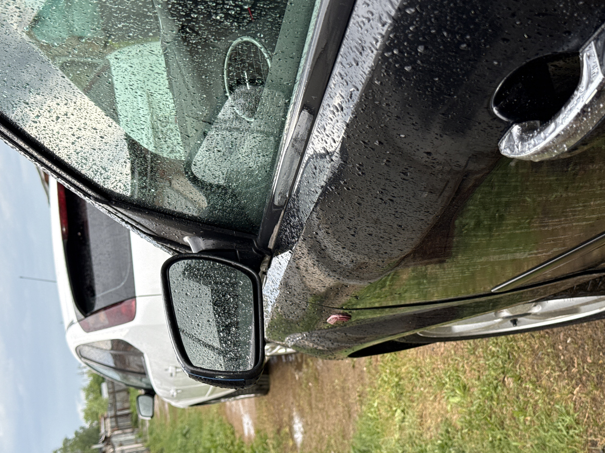 A close-up view of a black car, potentially parked in Chaikovsky, Russia. Raindrops cover the car's surface and windows. A white car is visible behind it, reflected in the side mirror. The ground alongside the cars has green grass.