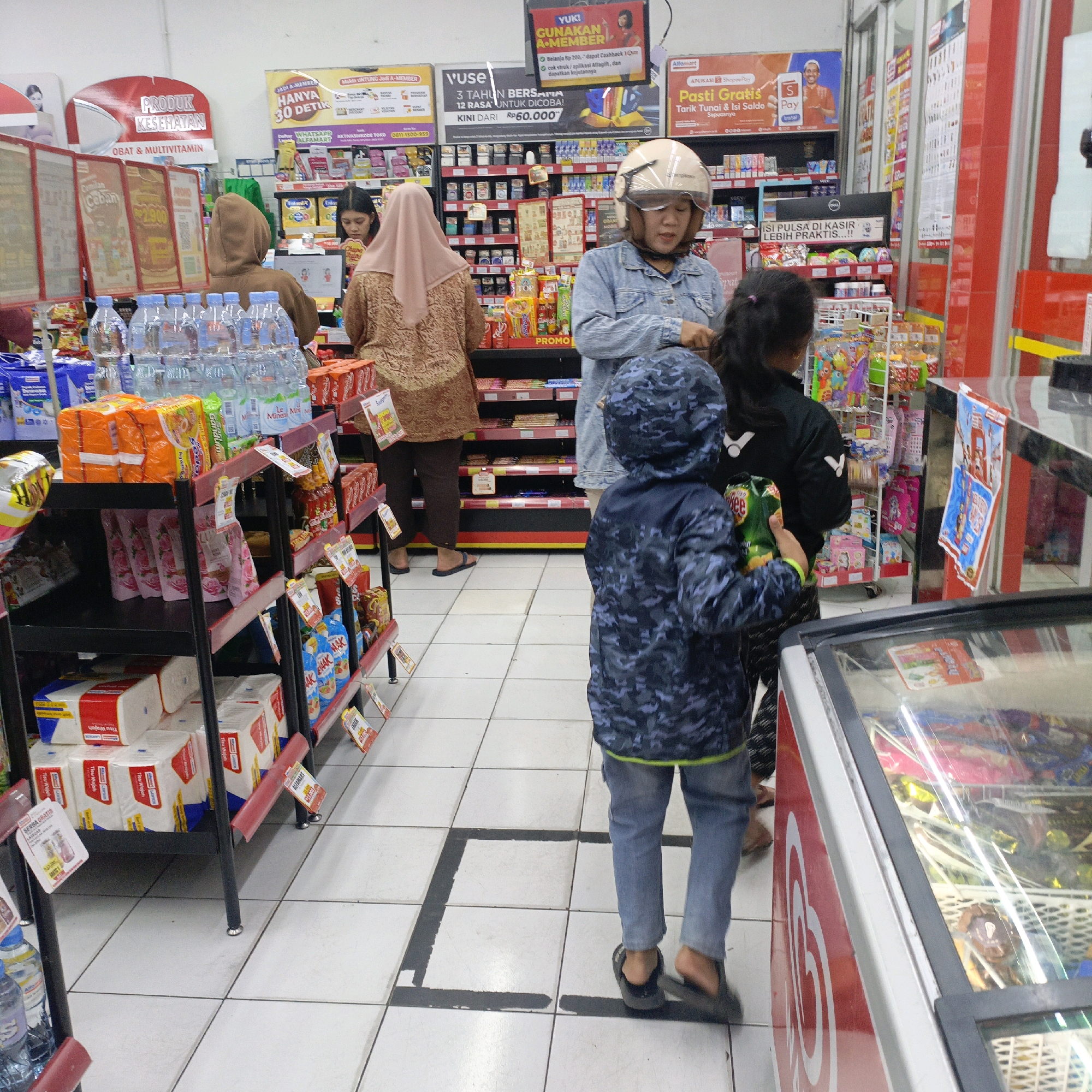 The image depicts the interior of a minimarket, specifically an Alfamart store, located in Salatiga, Indonesia. The perspective is from a low angle, looking towards the checkout area and a shelf stocked with various products.
In the foreground, a child wearing a dark blue camouflage jacket and light blue jeans stands facing away from the camera, holding a green bag of snacks. To the right, a glass-fronted cooler displays an assortment of frozen treats, including ice cream. A red and white branded sign is visible on the side of the cooler.
In the middle ground, several people are positioned near the checkout counter. A woman wearing a helmet and a light blue denim jacket is interacting with the cashier, whose face is not clearly visible. Another woman, dressed in a brown headscarf and a patterned brown dress, stands to the left, also appearing to be interacting with the checkout process. A third woman, partially obscured, stands further back, wearing a light pink headscarf and a brown patterned garment.
Behind the people, shelves are densely packed with a variety of consumer goods, including bottled water, packaged snacks, cleaning supplies, and health and beauty products. Promotional posters and signage are visible on the walls and shelves, advertising various offers and brands. The floor is tiled in a white and grey checkered pattern, with a black painted rectangle on the floor indicating a queueing area. The overall environment suggests a typical retail setting in Indonesia.
